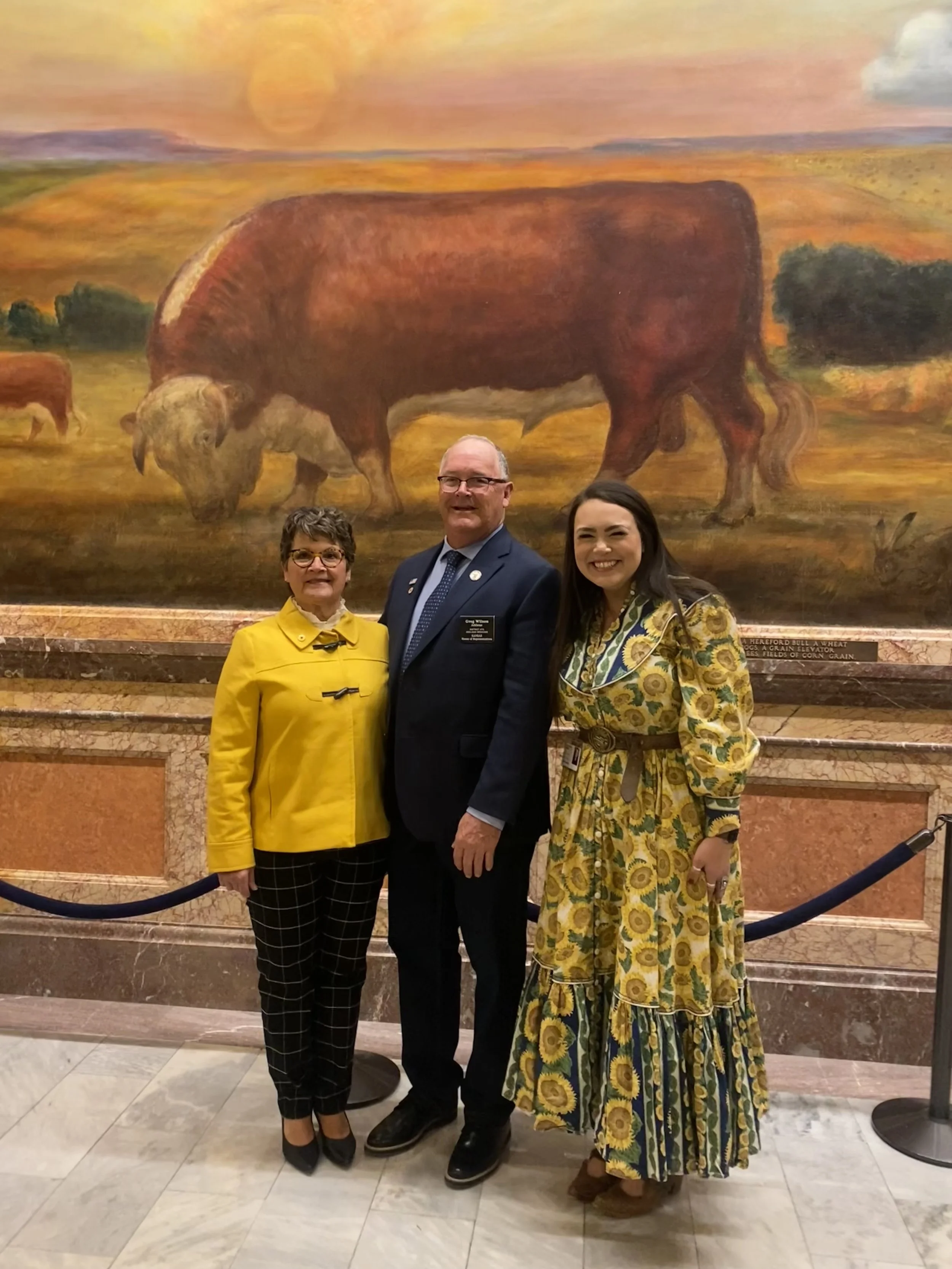 Three people standing in front of a large painted landscape of cattle grazing in a field at sunset, with a marble and stone wall and velvet rope barrier in the foreground.