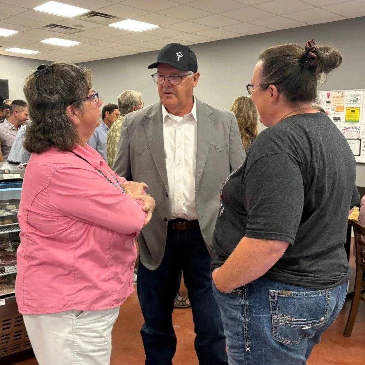 Three people engaged in a conversation in a crowded indoor setting, including an older man in a gray blazer and black cap, a woman in a pink jacket, and a woman in a dark t-shirt with glasses.