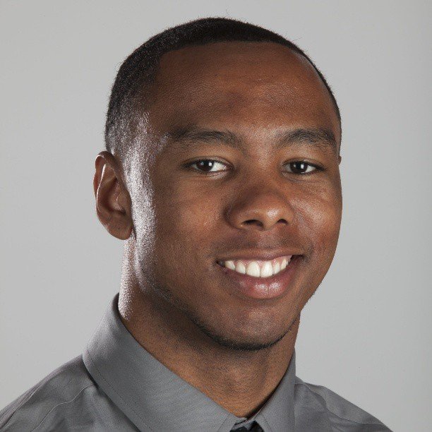 A young man with short black hair, smiling, wearing a gray collared shirt against a plain light gray background.
