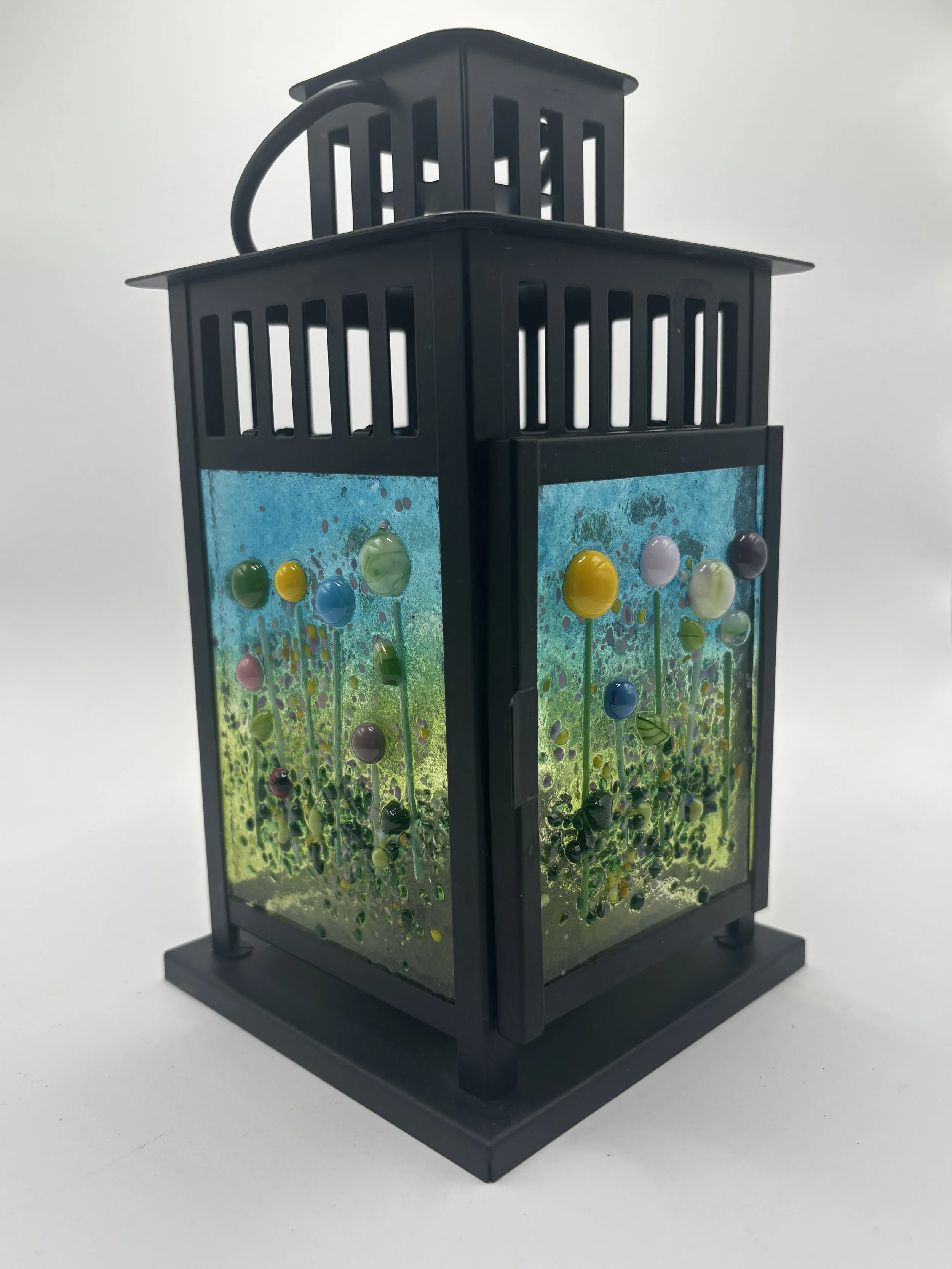 A decorative lantern with a black frame, colorful glass flowers, and a painted background with green, yellow, blue, purple, and black spots, placed on a plain white surface.