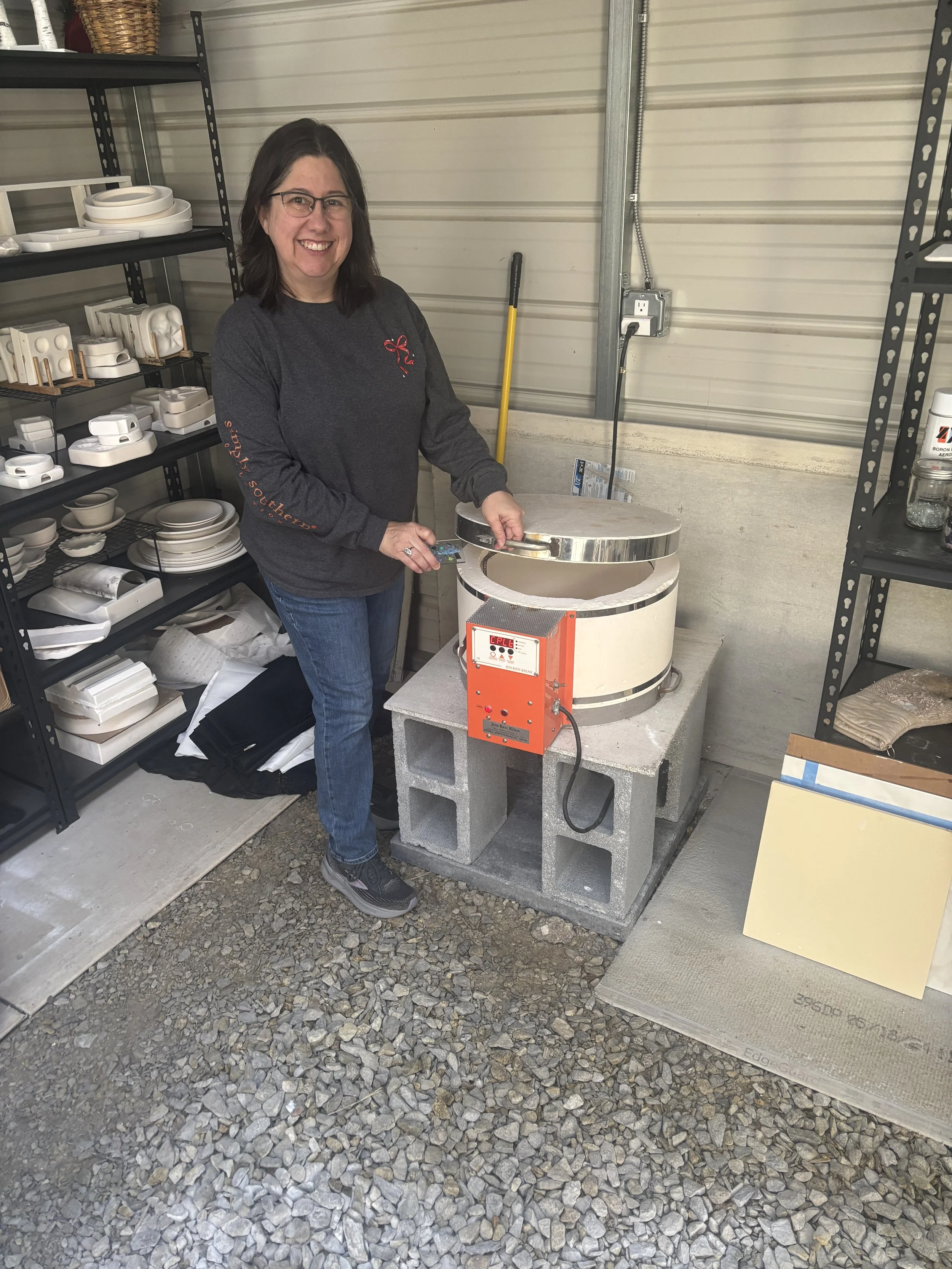 A woman in a gray long-sleeve shirt with glasses is smiling and working with a kiln on cinder blocks in a garage or workshop. Shelves with white ceramics and pottery items are visible to her left.
