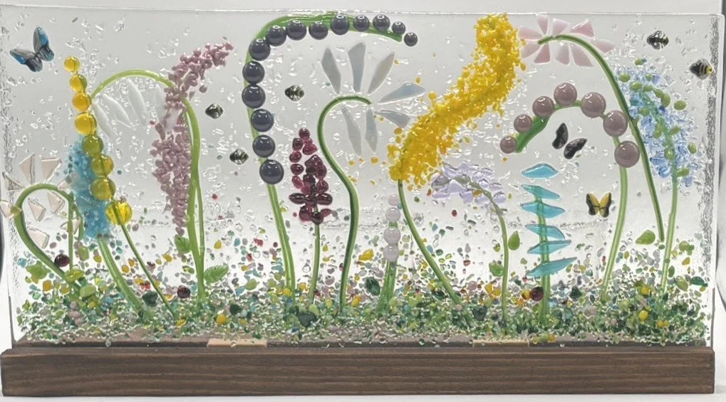 Colorful decorative glass art featuring various flowers and butterflies, with a wooden base and a background filled with tiny bubbles.