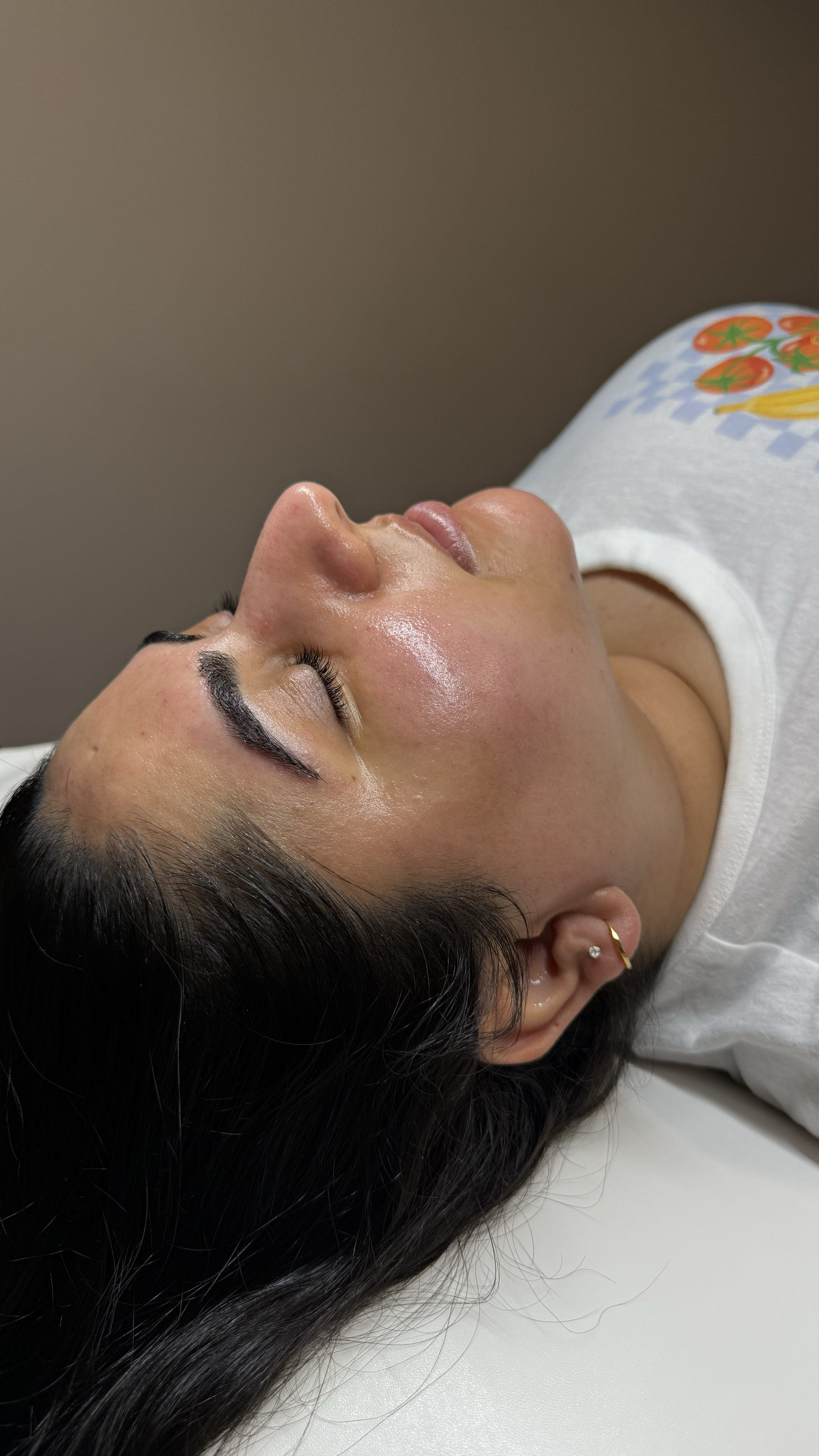 A woman with dark hair and earrings is receiving a facial treatment, lying down with her eyes closed, in a spa or clinic setting.