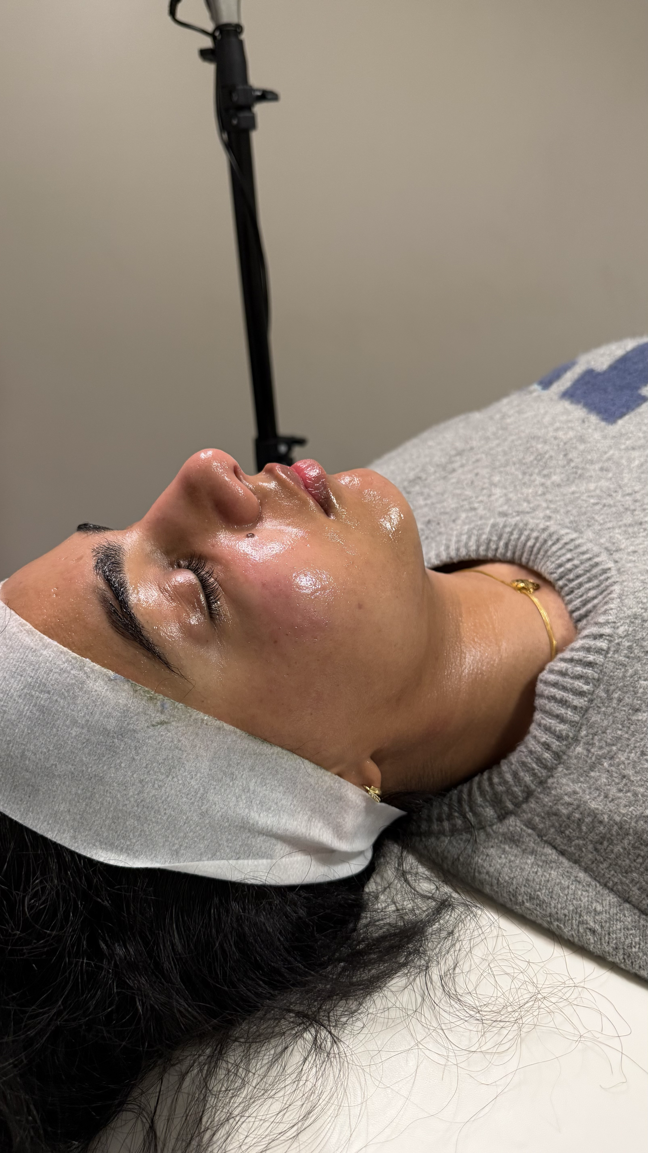 A woman lying down with eyes closed, wearing a white headband and gray sweater, appears to be receiving a facial treatment in a studio or clinic setting.