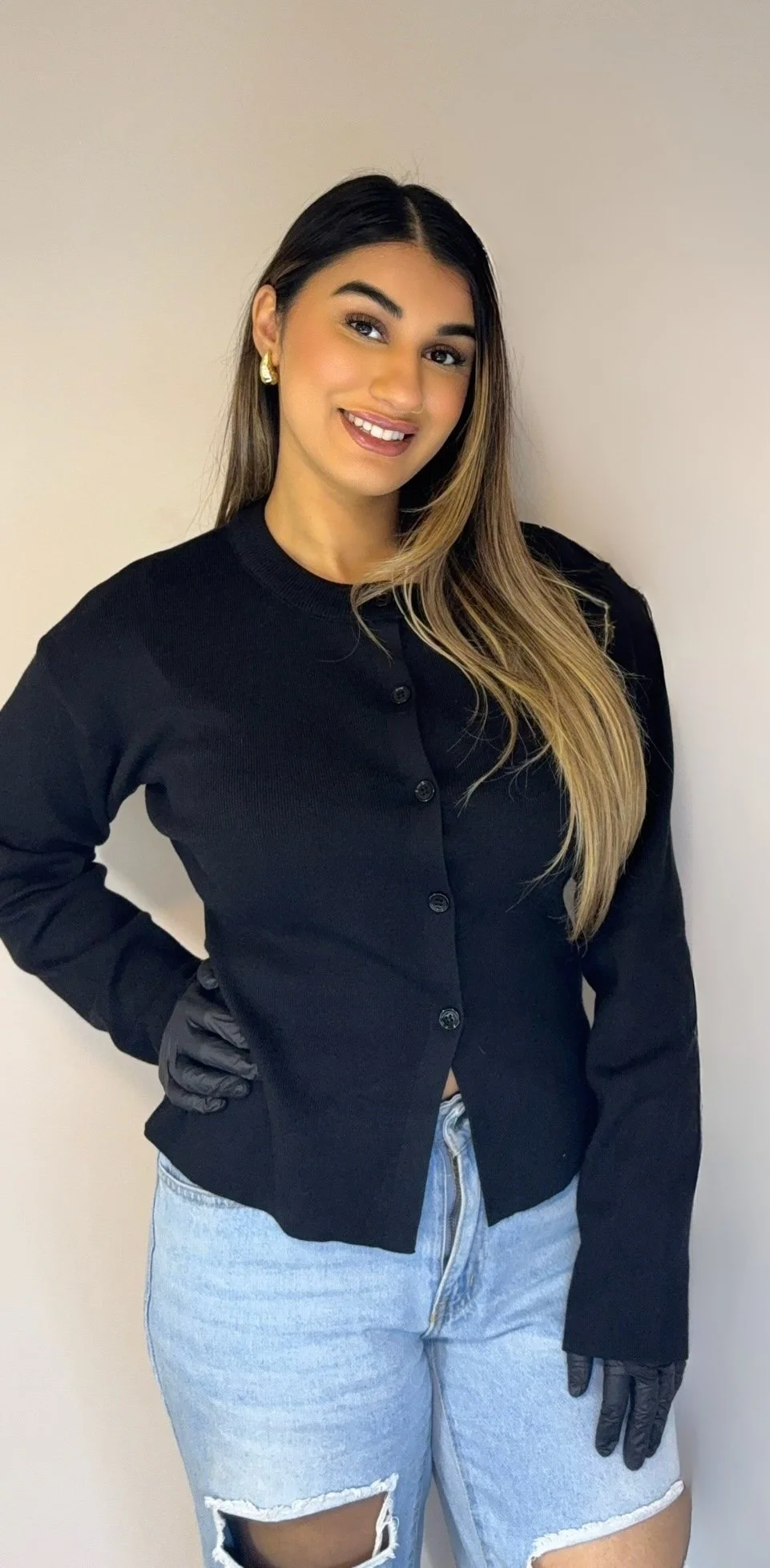 A young woman with long, wavy hair, wearing a black cardigan, light blue ripped jeans, and black gloves, standing against a plain beige wall, smiling at the camera.