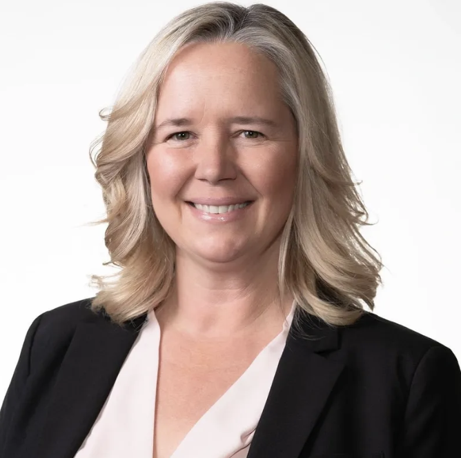 A professional woman with blonde, wavy hair wearing a black blazer and white blouse, smiling against a white background.