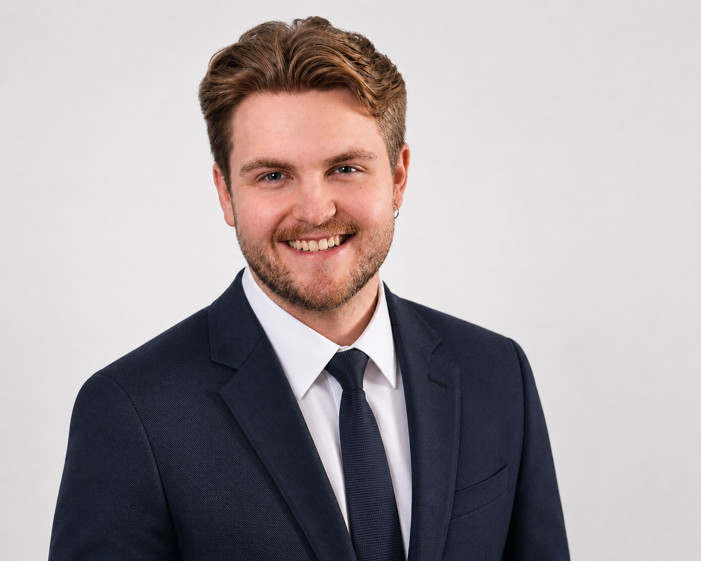 Professional headshot of a young man with styled light brown hair, light beard, wearing a dark suit, white shirt, and navy tie, against a plain gray background.