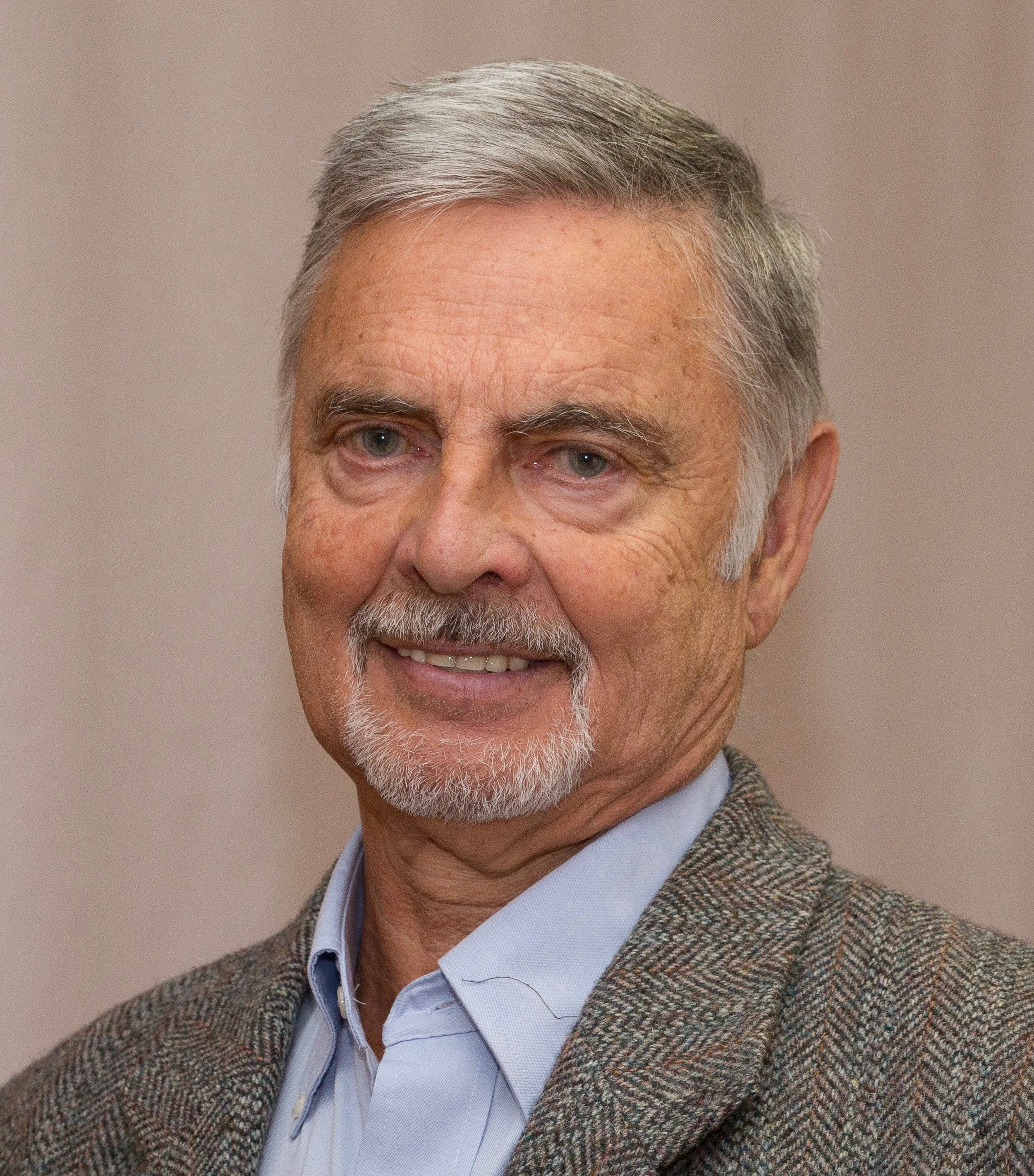 Close-up of senior man with gray hair and beard, wearing a tweed jacket and a collared shirt, smiling against a plain background.