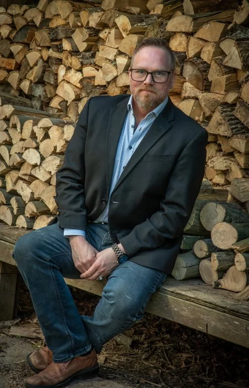 A man with glasses, a beard, and a mustache, dressed in a black blazer, light blue shirt, and jeans, sitting on a wooden bench in front of a large stack of firewood.