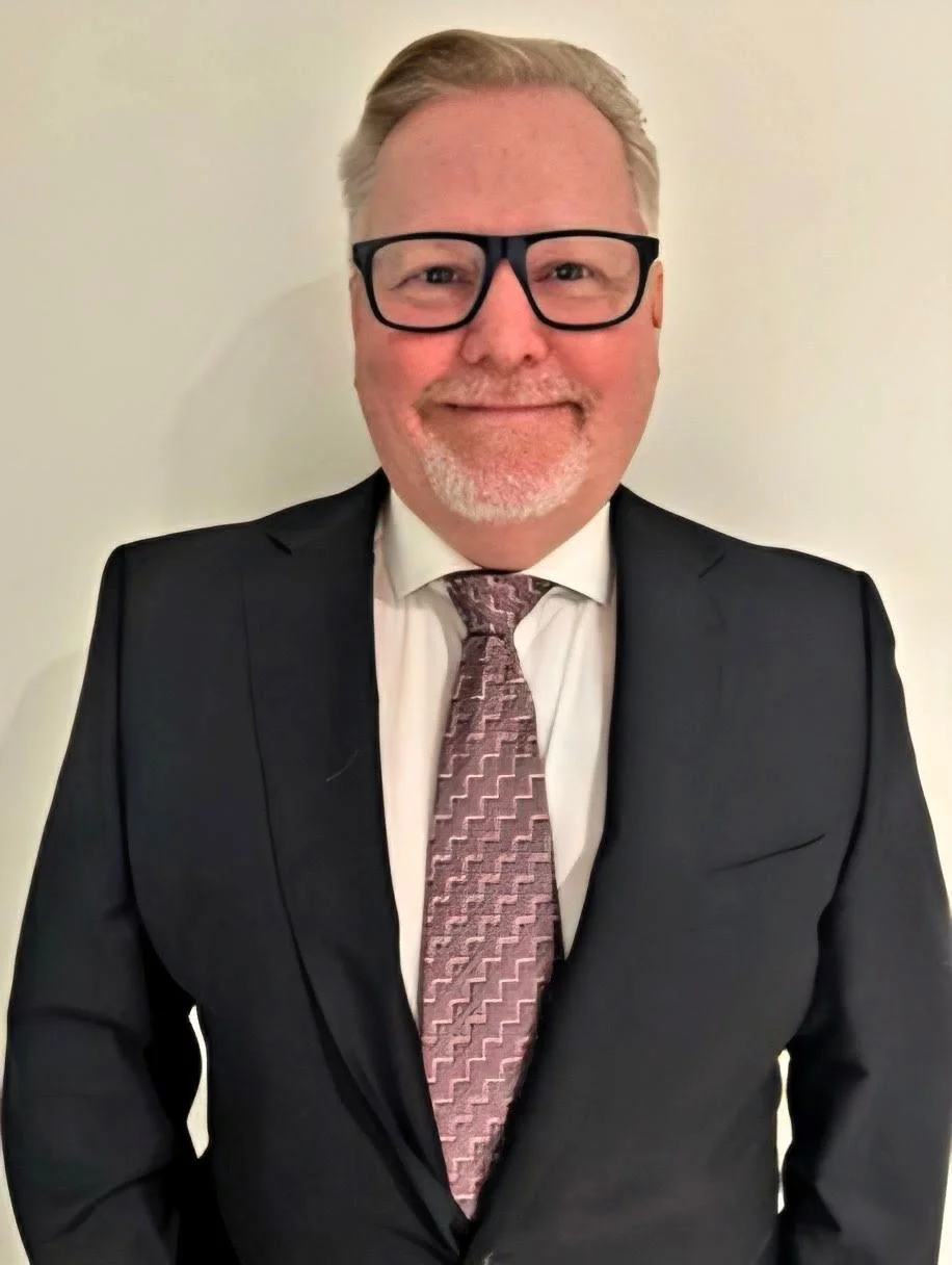 A middle-aged man with glasses, a light beard, and short light-colored hair, wearing a black suit, white shirt, and patterned tie, smiling in front of a plain off-white background.
