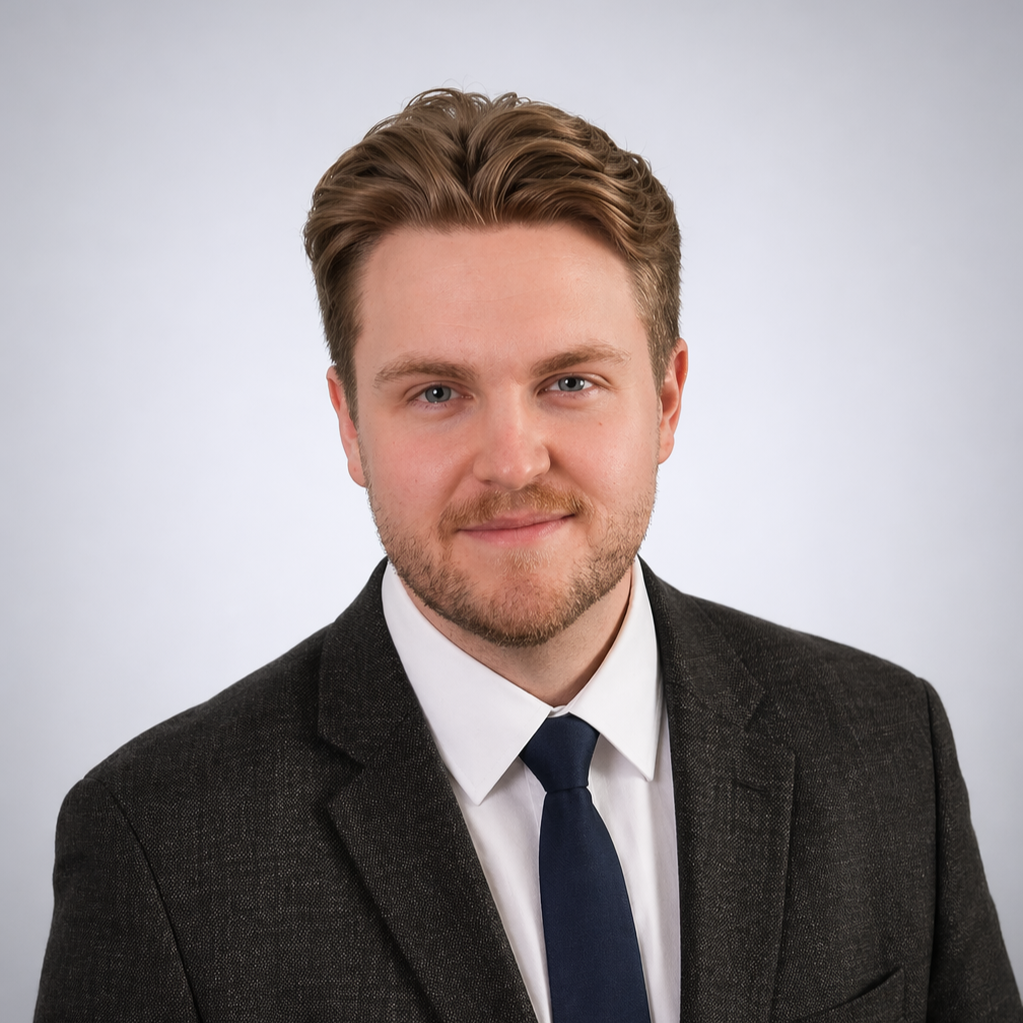 Professional headshot of a young man with styled light brown hair, light beard, wearing a dark suit, white shirt, and navy tie, against a plain gray background.