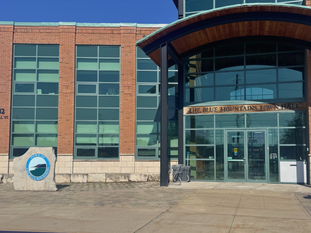 Exterior view of The Blue Mountains Town Hall building with a glass front entrance, a sign above the door, a stone with the town seal, and a shopping cart near the entrance.