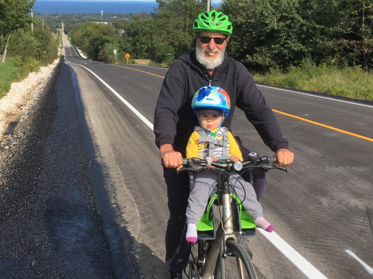 A man and a young child riding bikes on a countryside road with trees and hills in the background. The man is wearing sunglasses, a black hoodie, and a green helmet, while the child is in a bike seat wearing a blue helmet and yellow jacket.