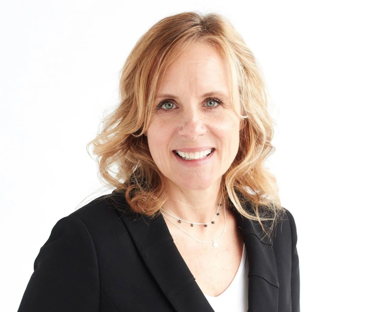 Smiling woman with blonde curly hair wearing a black blazer and layered necklaces, against a white background.