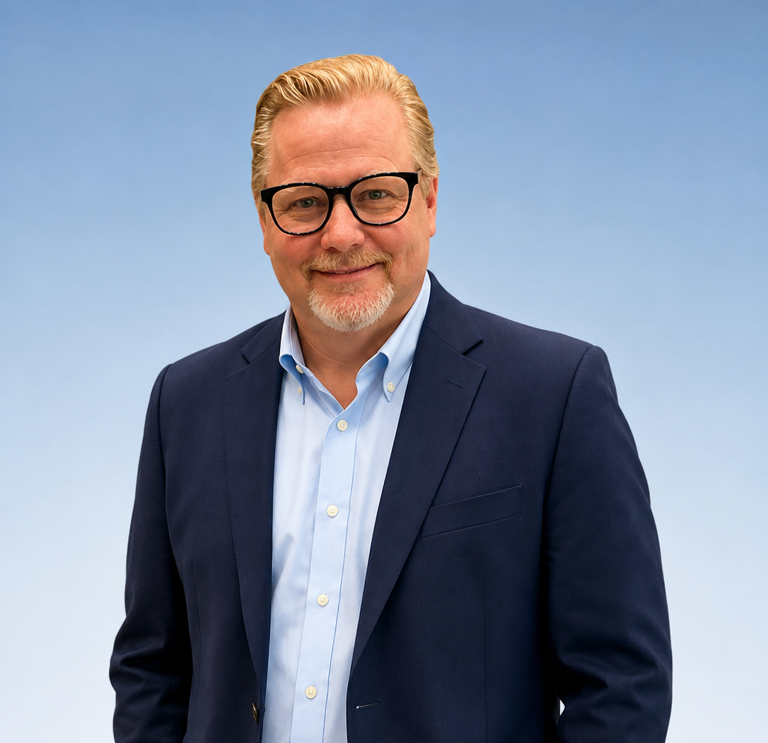 A middle-aged man with glasses, a light beard, and short light-colored hair, wearing a black suit, white shirt, and patterned tie, smiling in front of a plain off-white background.