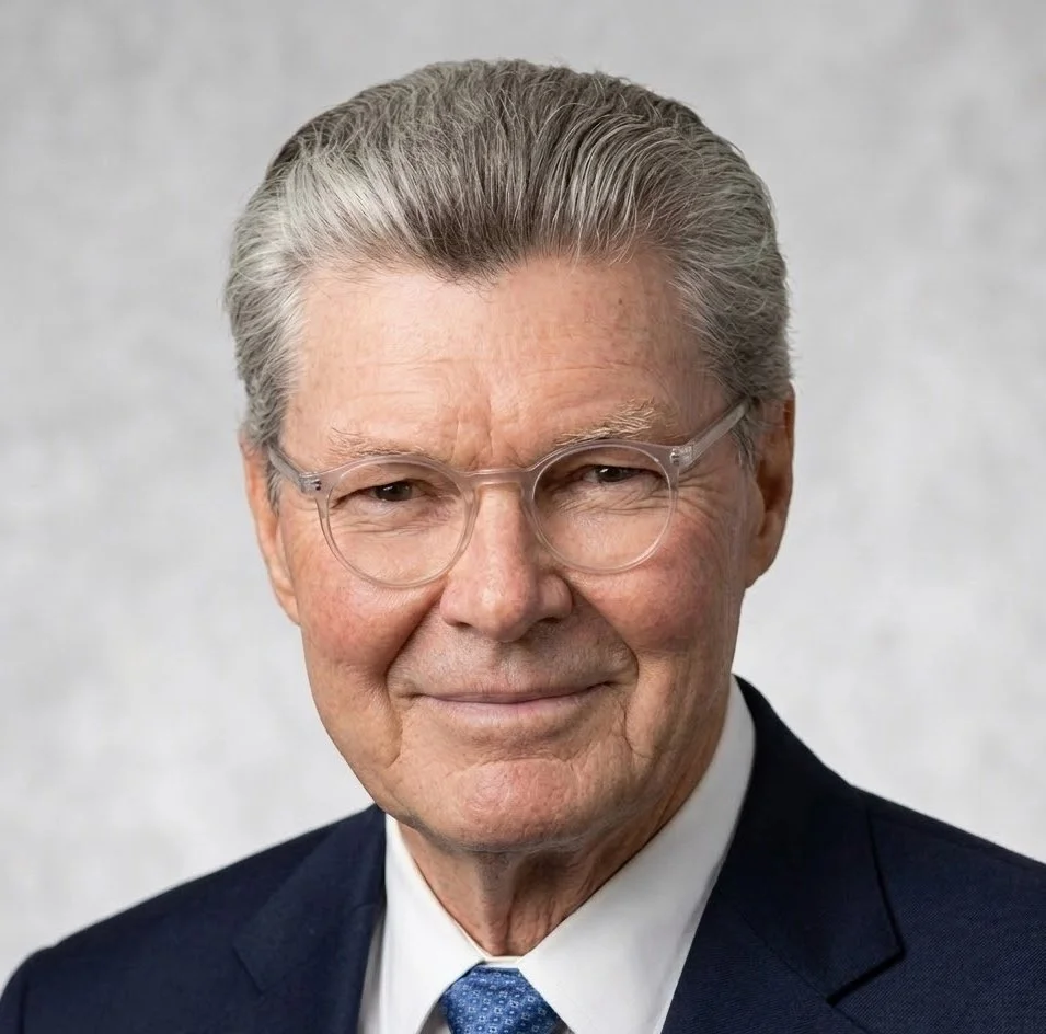 Close-up portrait of an elderly man with gray hair, glasses, wearing a dark suit, white shirt, and blue tie, smiling gently against a light gray background.