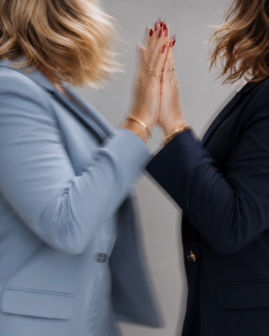 Two women in business suits touching foreheads and pressing their hands together in a gesture of solidarity or greeting.