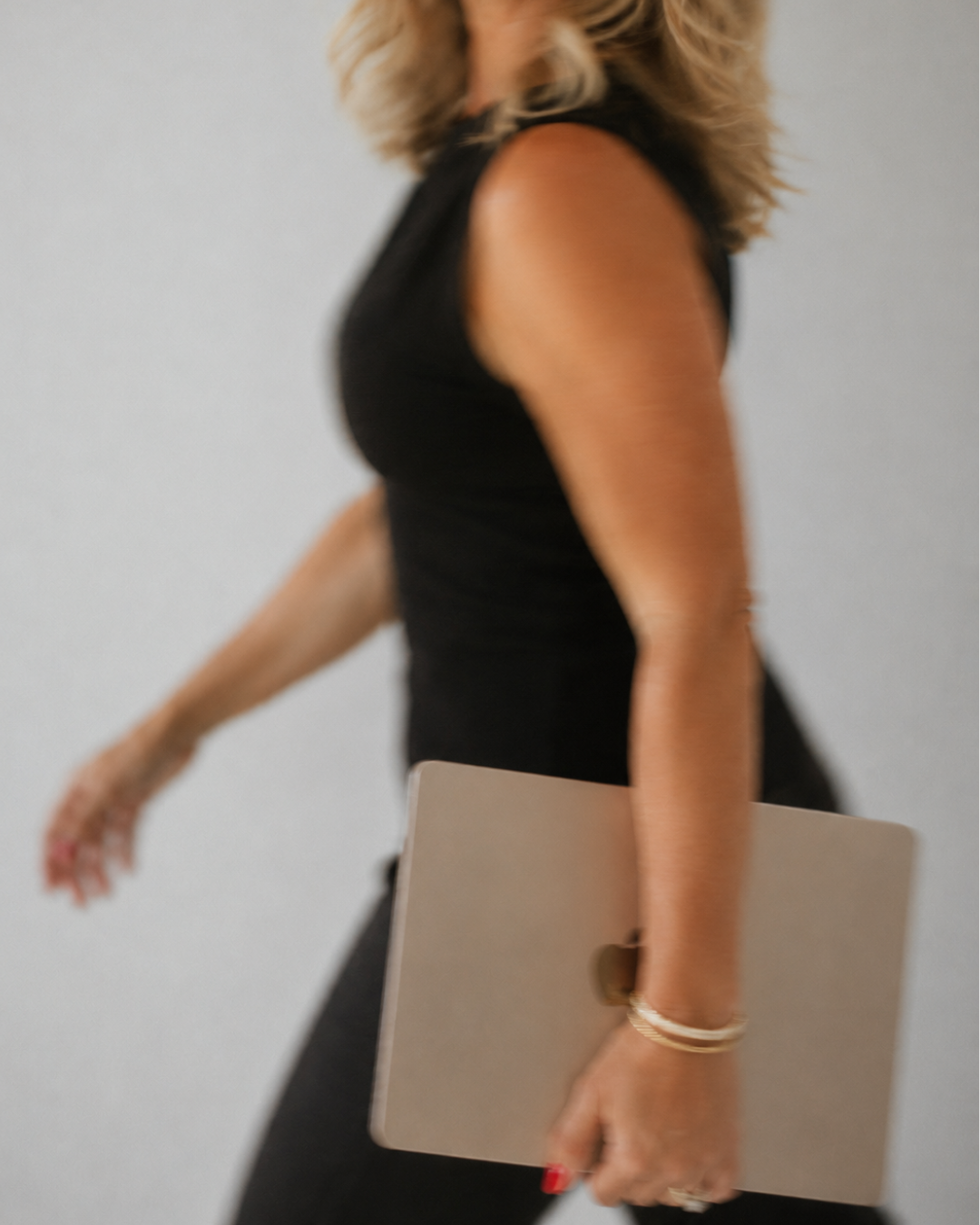 A woman dressed in black holding a closed laptop, with her face not visible, against a plain background.
