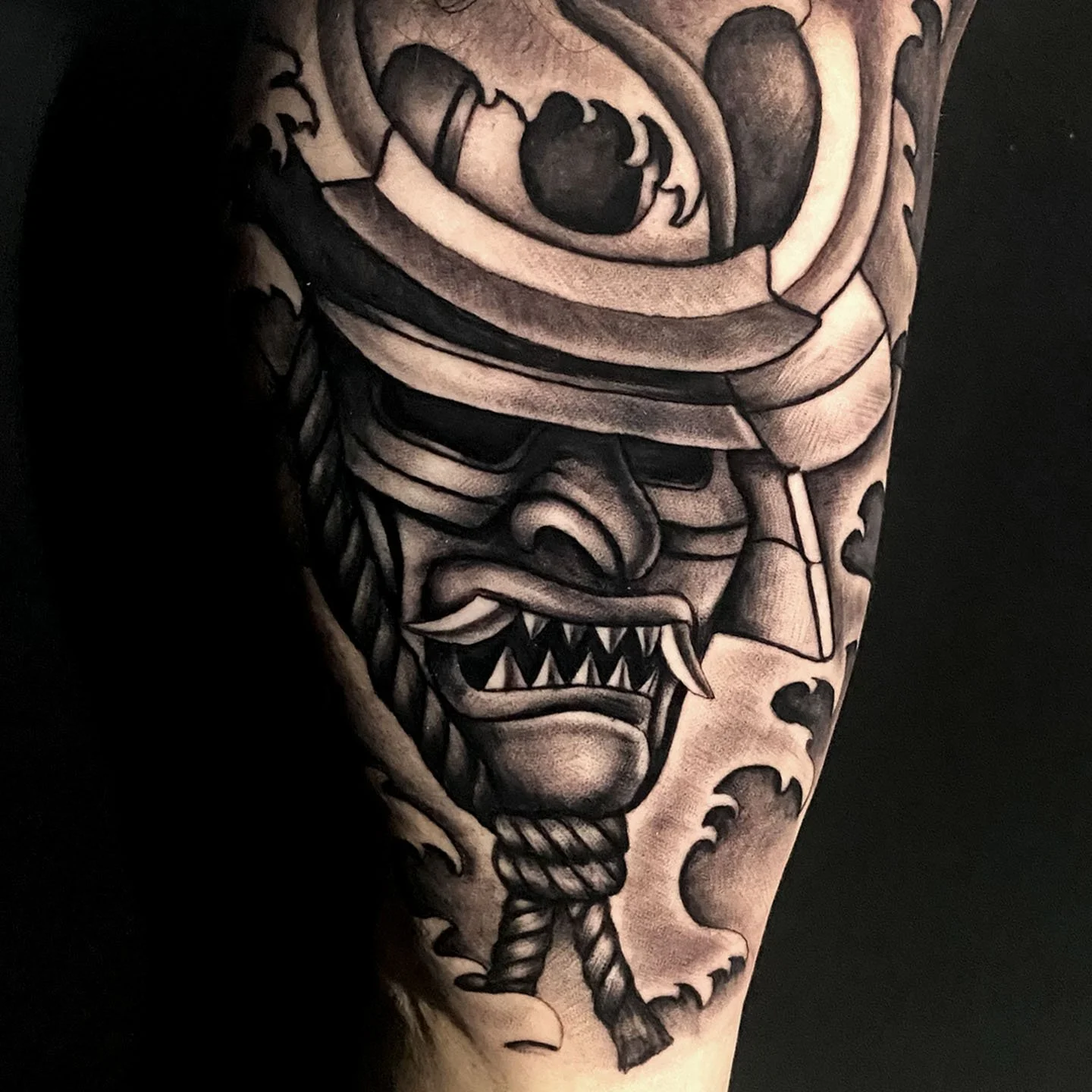 Black and gray tattoo of a fierce, stylized face with sharp teeth, an eyepatch, and a bandana, on a person's upper arm.
