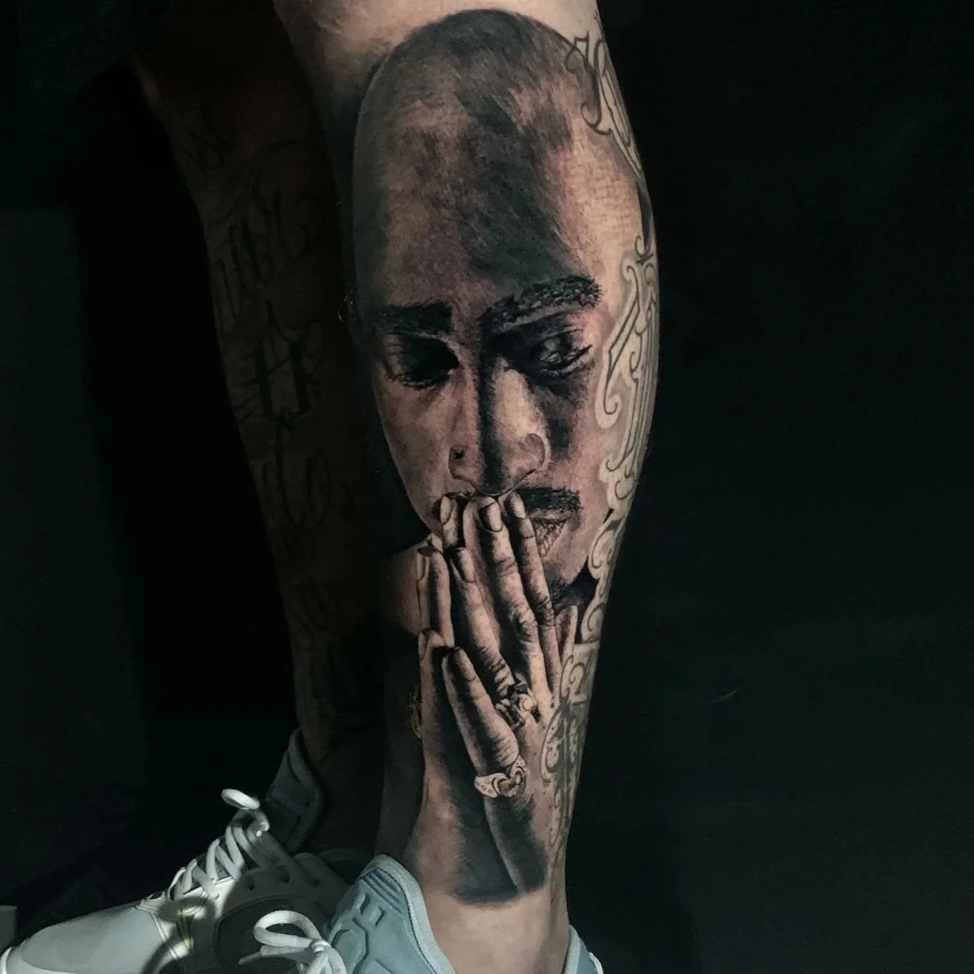 Tattooed arm of a person with a detailed black and gray portrait of a person with their hands in prayer position, tattoos on the arm and hand, wearing white sneakers.