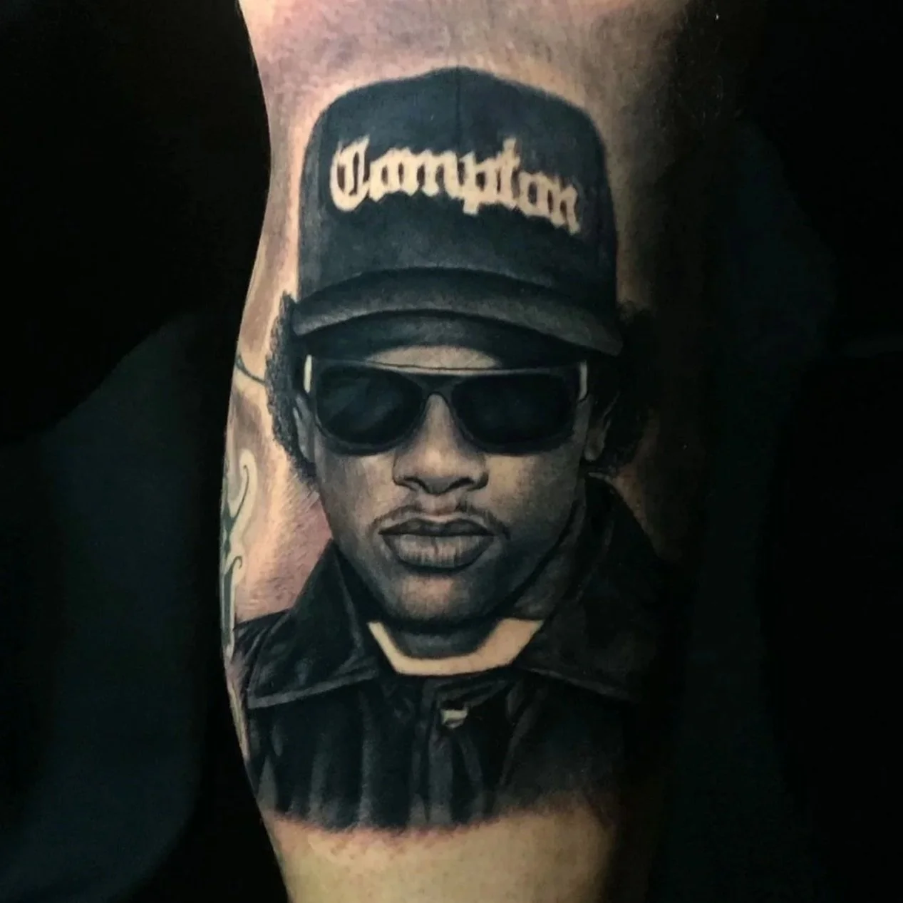 Black and gray tattoo of a man's face wearing sunglasses and a cap with the word 'Dantoni' on it, on a person's arm.