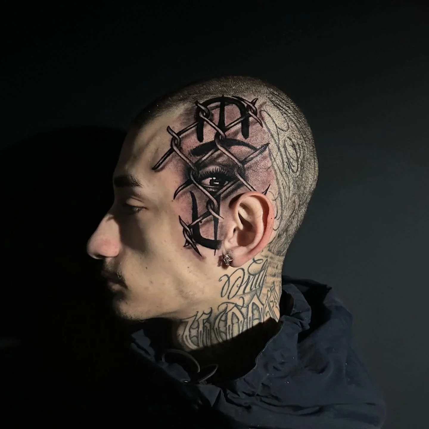 Side profile of a person with facial tattoos, shaved head, and an ear piercing, against a dark background.