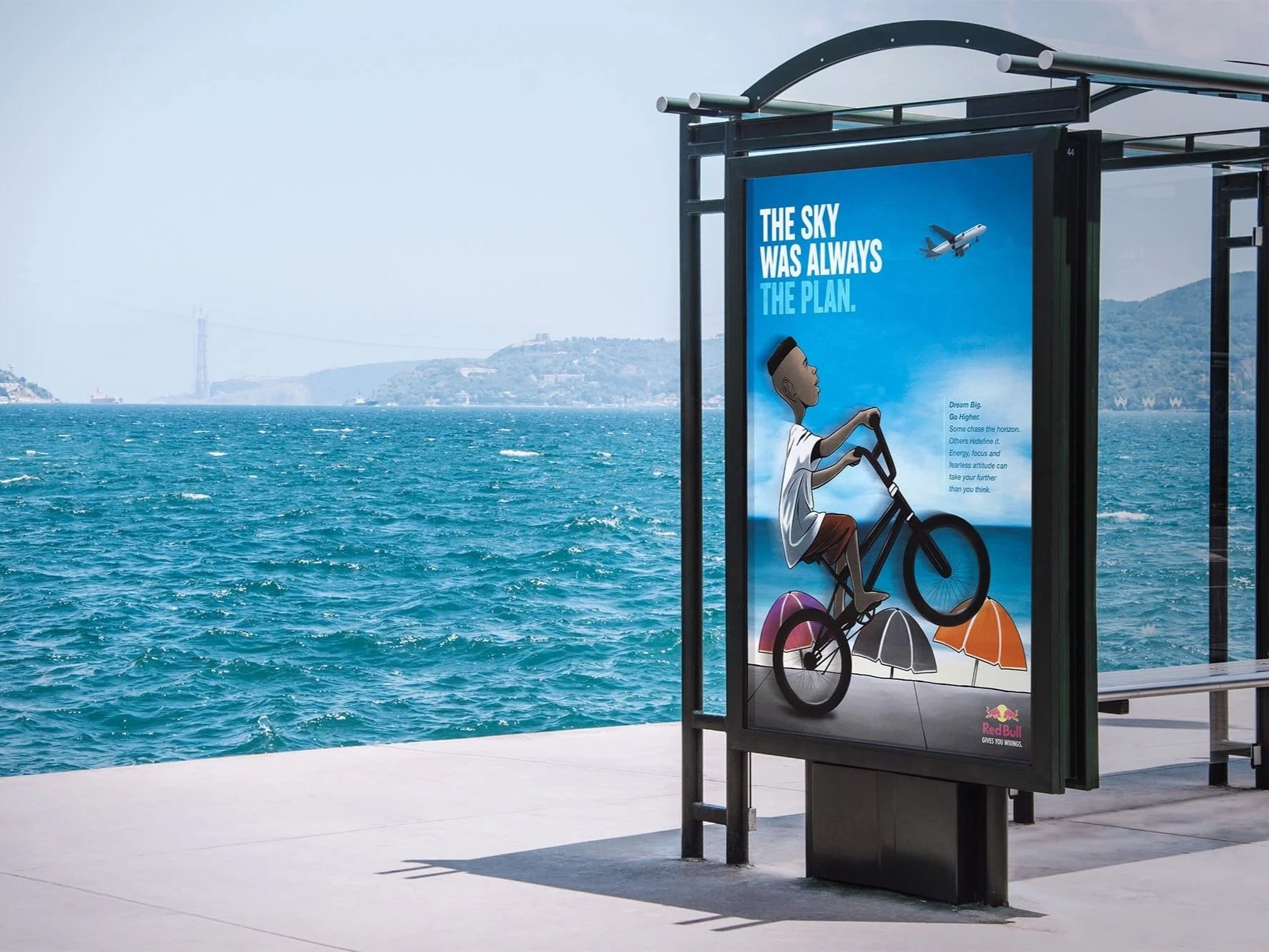 An outdoor bus stop advertisement showing a cartoon boy riding a bicycle with beaches and ocean in the background. The ad promotes dreaming big and encourages going higher, with a plane flying in the sky. There is also a Red Bull logo at the bottom.