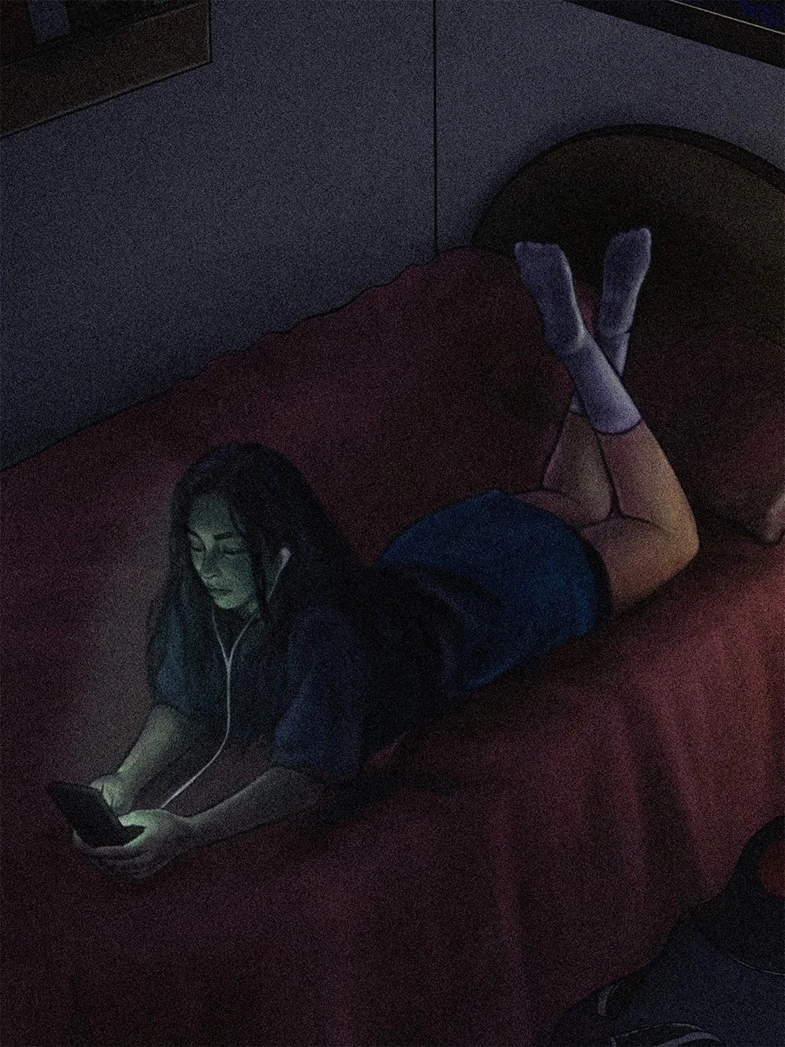 A woman lying on her stomach on a bed, wearing headphones and looking at her phone in a dimly lit room.