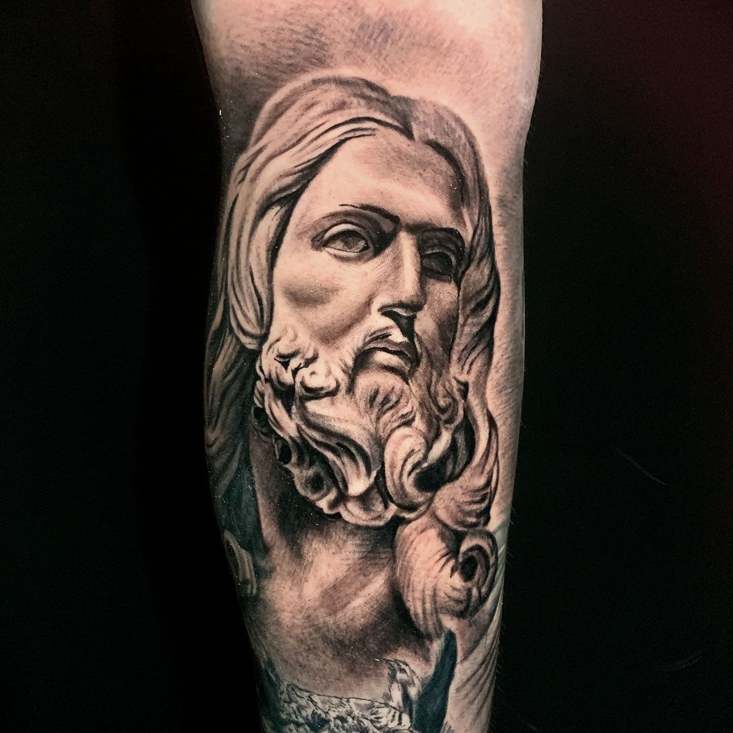 Black and gray tattoo of a detailed portrait of a bearded man with flowing hair, likely a depiction of Jesus Christ, on a person's arm.