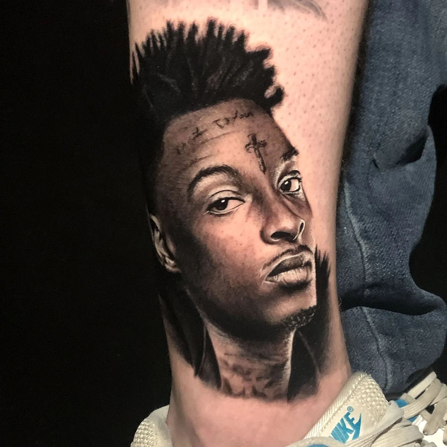 A detailed tattoo of a person's face with short dreadlocks, tattoos on the forehead and neck, wearing a white Nike shoe.