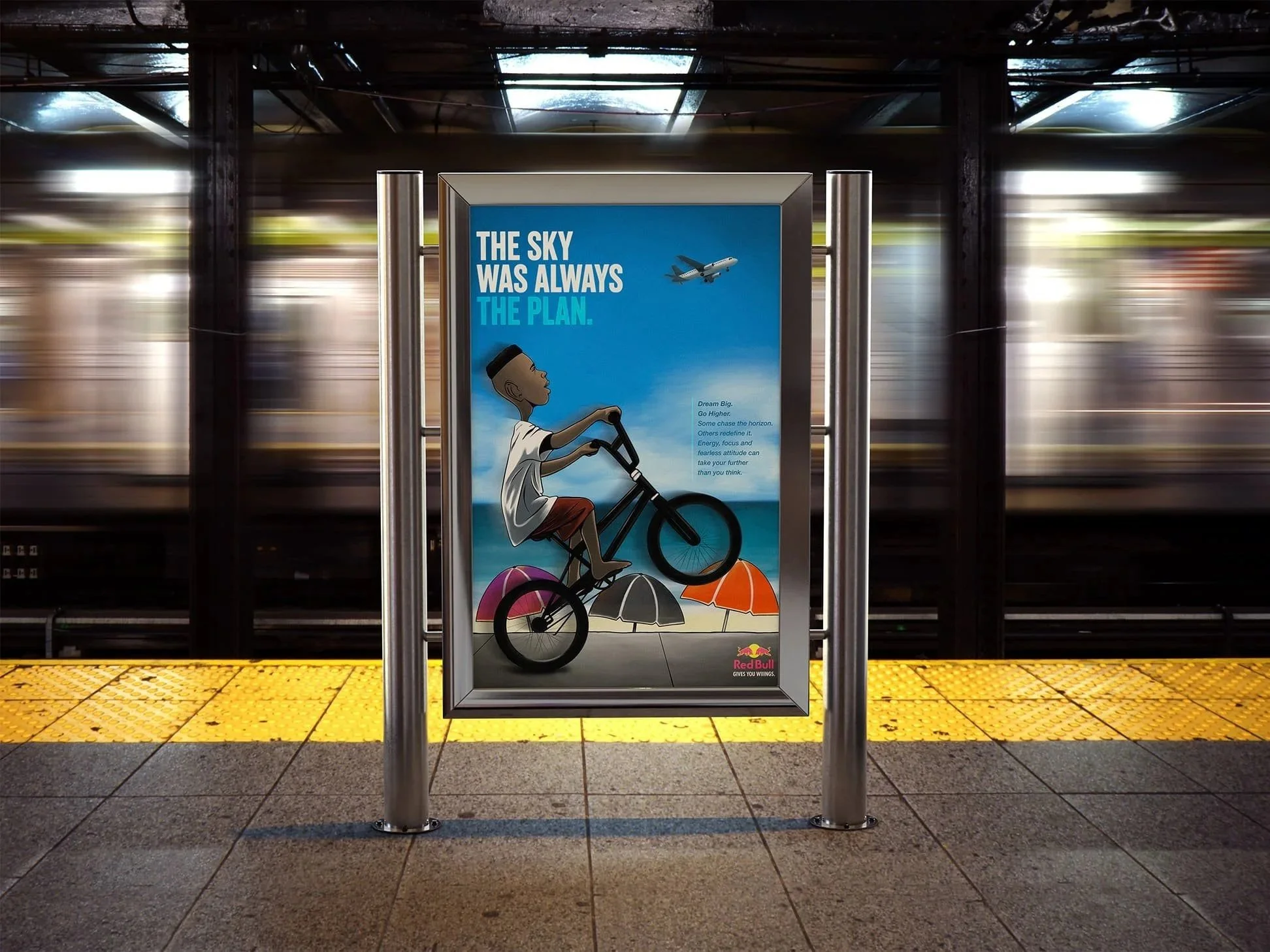 A poster on a subway platform shows a cartoon boy riding a bicycle on the beach with umbrellas and text that says, 'The sky was always the plan.' An airplane is flying in the sky in the poster. The Red Bull logo is at the bottom of the poster.