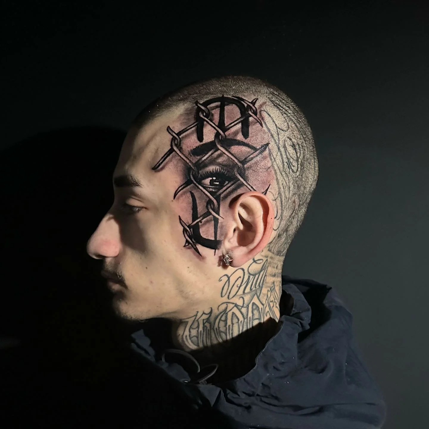 Side profile of a person with facial tattoos, shaved head, and an ear piercing, against a dark background.