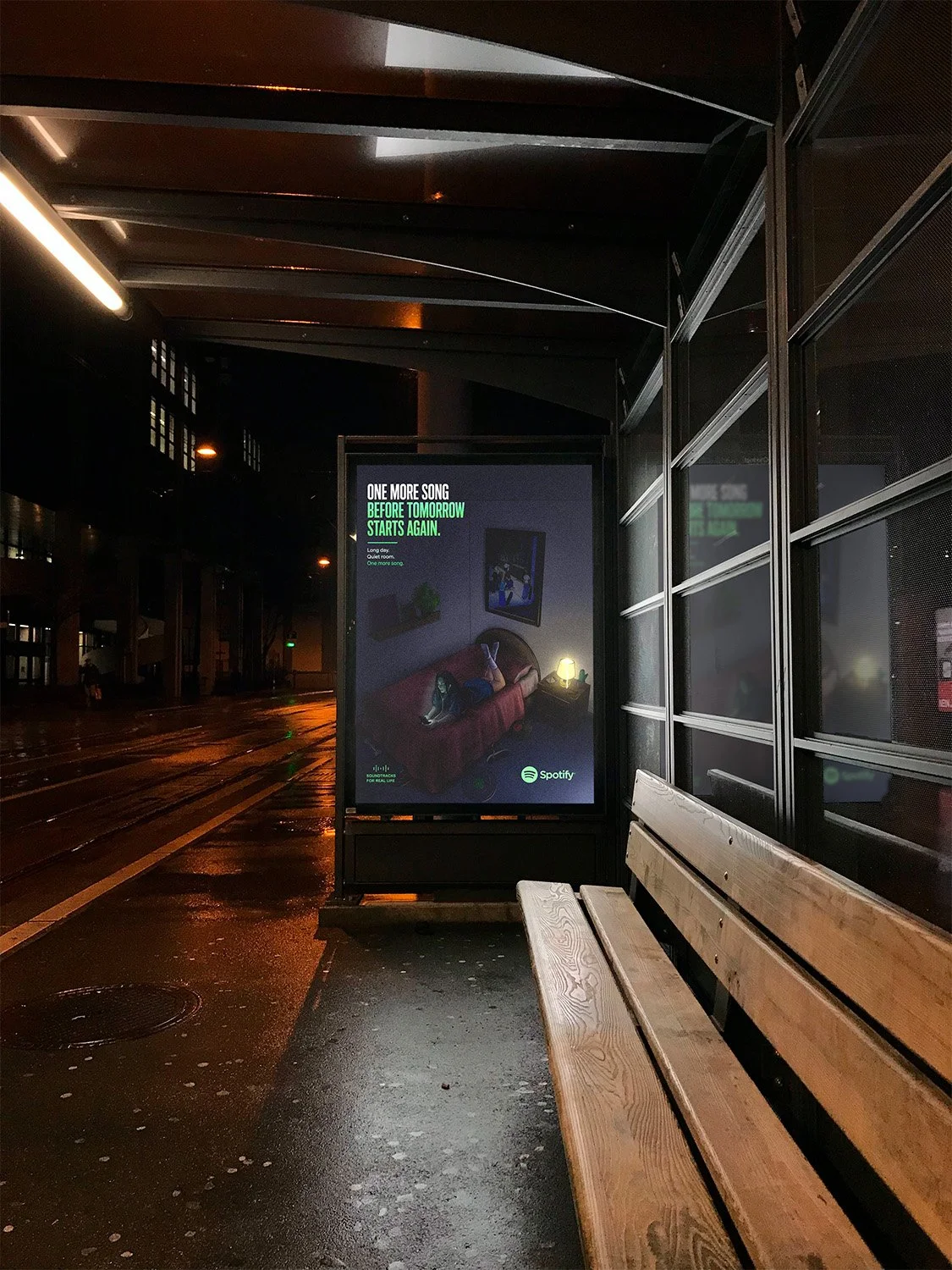Nighttime scene at a bus stop with a digital advertisement showing a person lying on a couch with headphones and a phone. The ad displays Spotify branding and the message: "One more song before tomorrow starts again." The bus stop has wooden benches, side panels, and a wet pavement reflecting nearby lights.