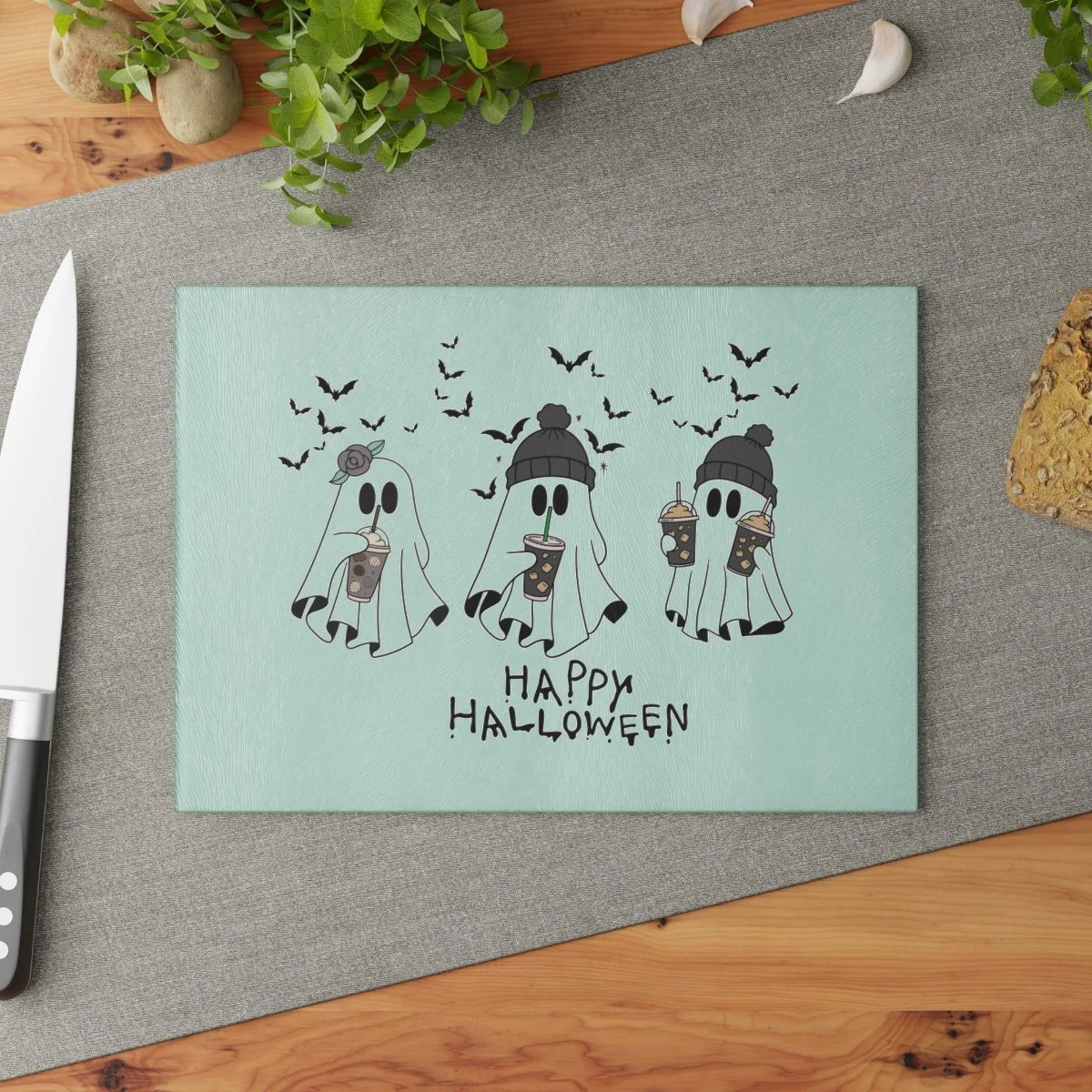Ghost Trio Cutting Board. Clear glass cutting board with 3 ghost in hats, holding coffees and bats flying in the background. The colors are black, white, and grey.. It also says "happy halloween".