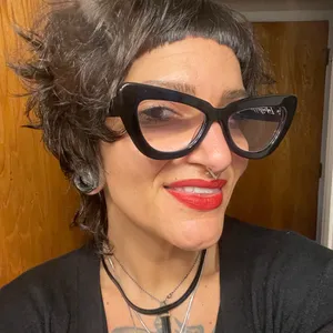 A woman with curly short dark hair and oversized black glasses, smiling with red lipstick, wearing layered necklaces, in front of a wooden background.