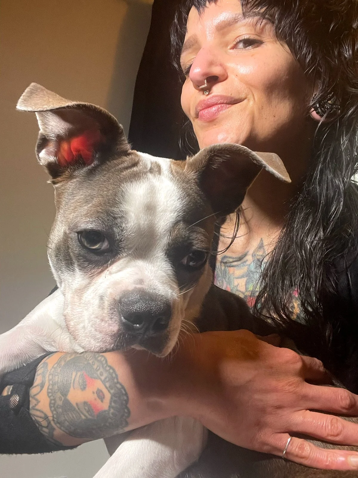 A person with tattoos and facial piercings holding a dog close to their face.