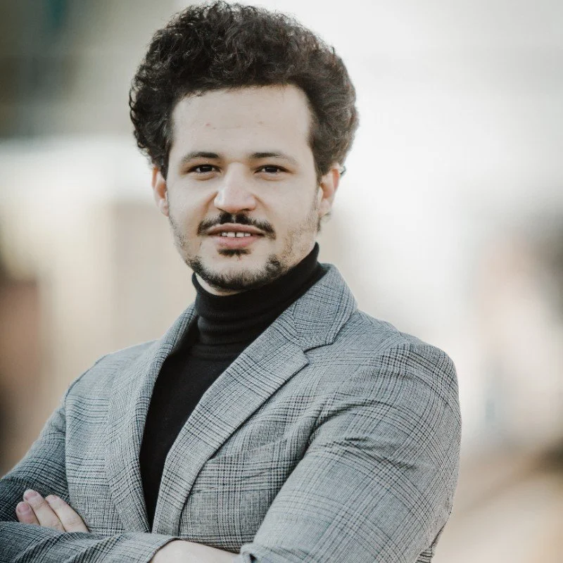 A man with curly dark hair and a beard, wearing a gray blazer and black turtleneck, standing outdoors with his arms crossed, smiling slightly.