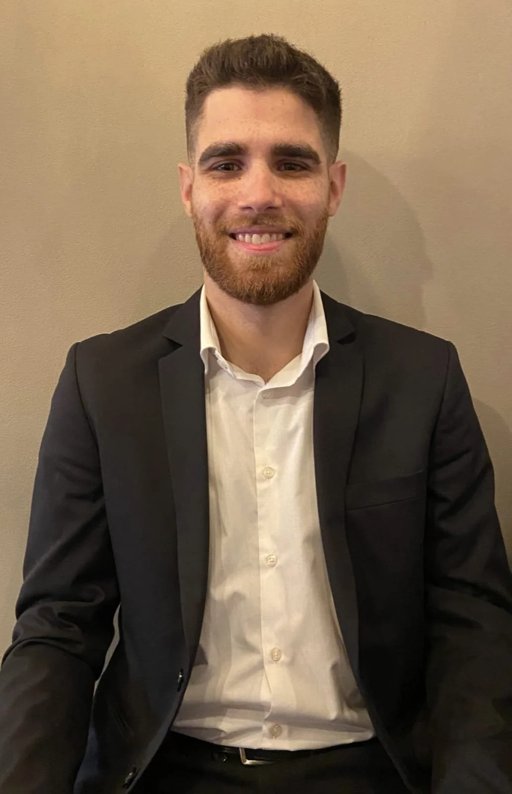 A young man with a beard and short brown hair, wearing a black suit jacket and white dress shirt, smiling and sitting against a beige wall.