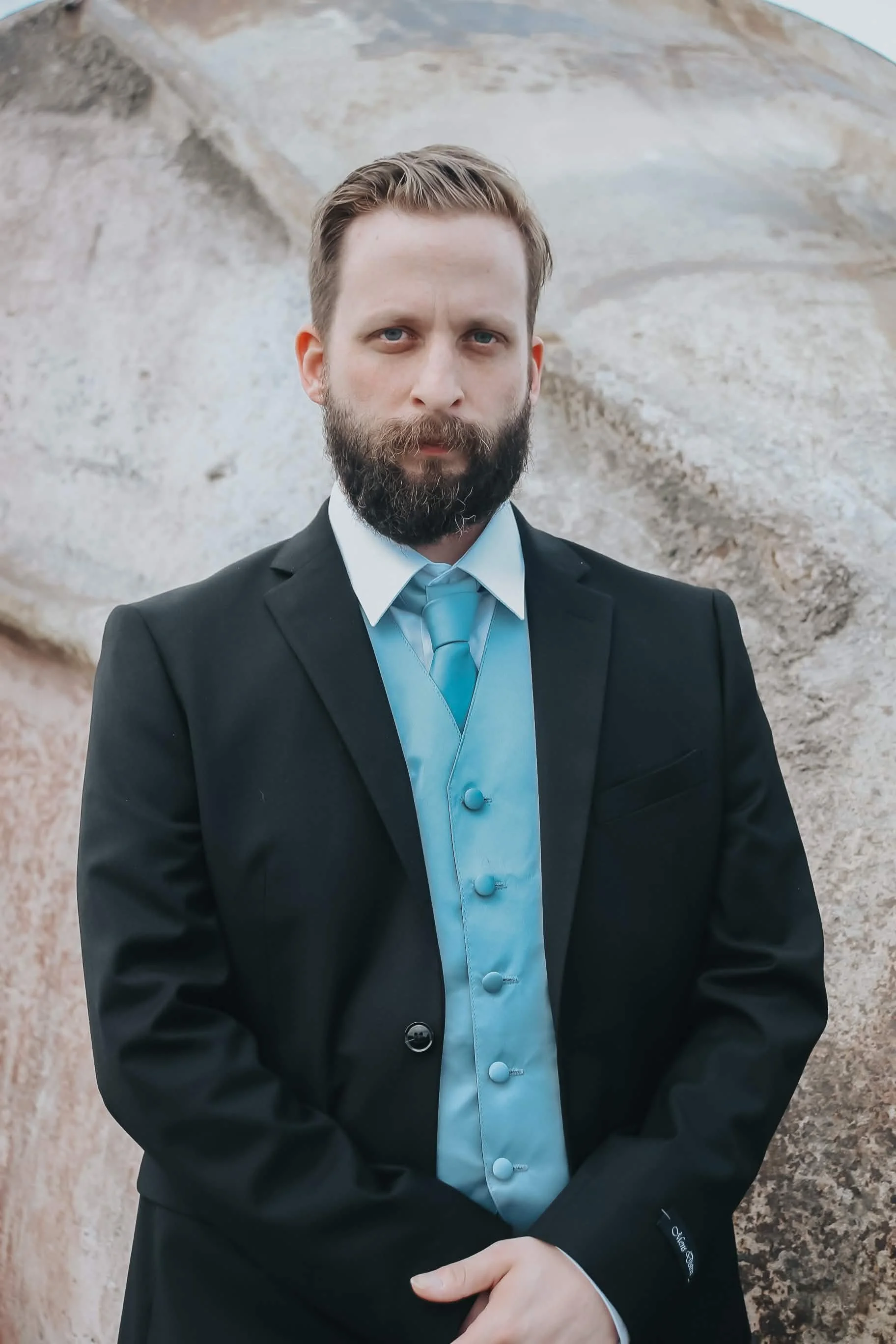 A man dressed in a black suit, light blue shirt with decorative buttons, and matching tie, standing in front of a rocky background. He has short, light brown hair and a full beard.