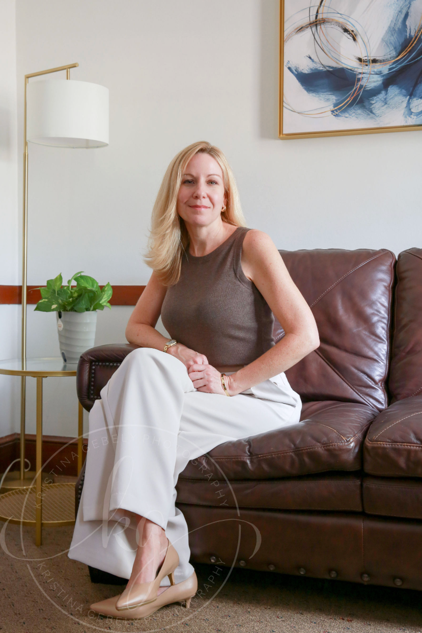 A woman sitting on a brown leather couch in a living room, wearing a sleeveless brown top and white pants, with a plant on a side table and an abstract painting on the wall behind her.