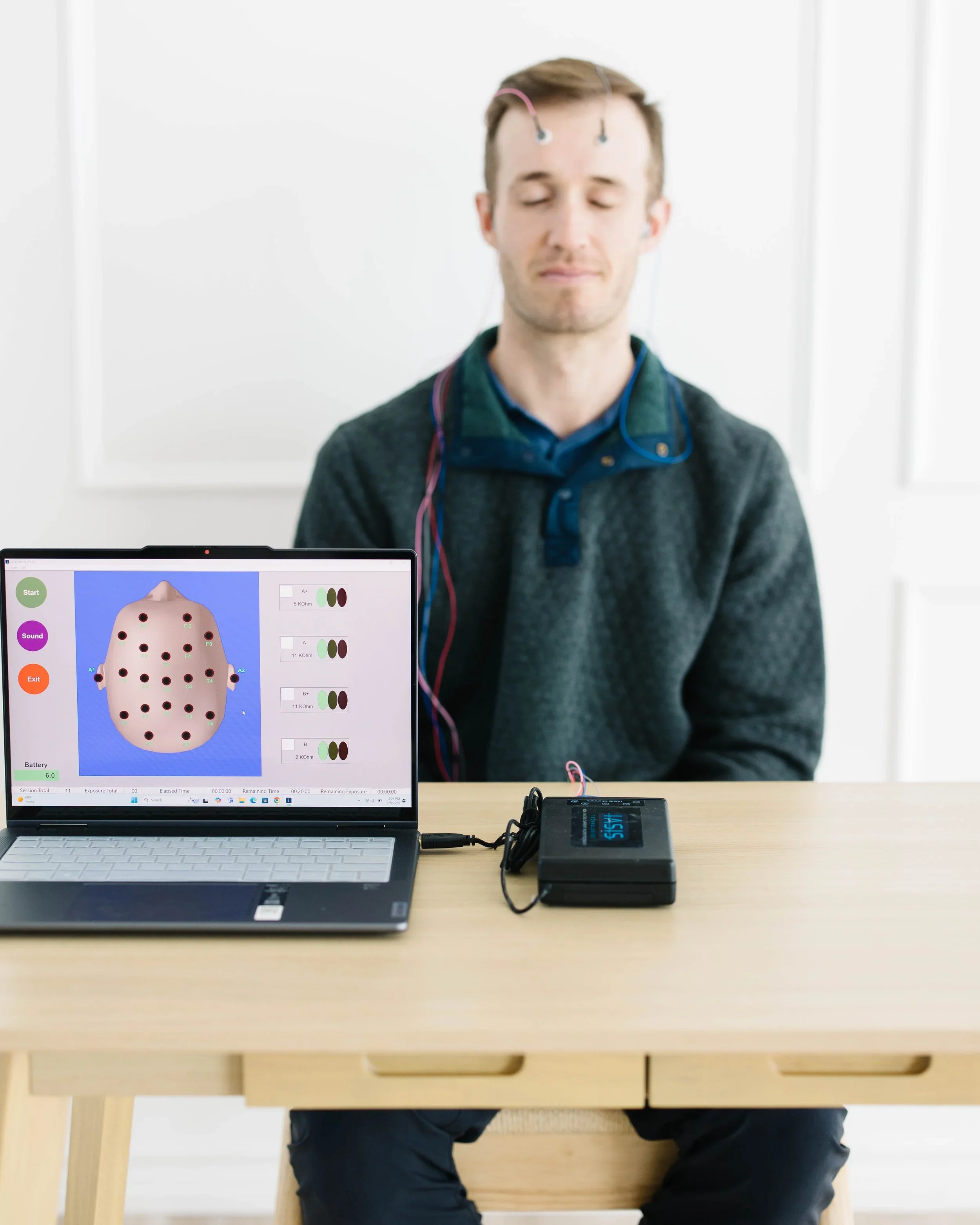 A man with electrodes on his head connected to wires sitting with eyes closed behind a computer displaying a diagram of the brain and EEG readings, in a clinical setting.