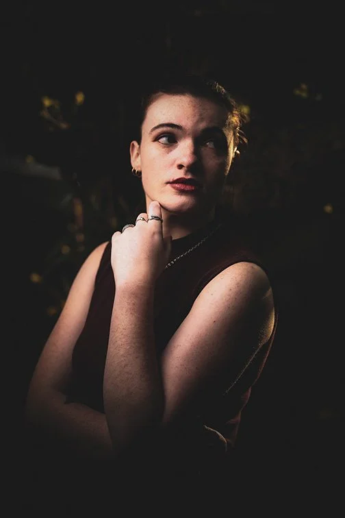 A young woman with dark hair and light skin, wearing a sleeveless top, touching her chin with her right hand, in a dark outdoor setting.