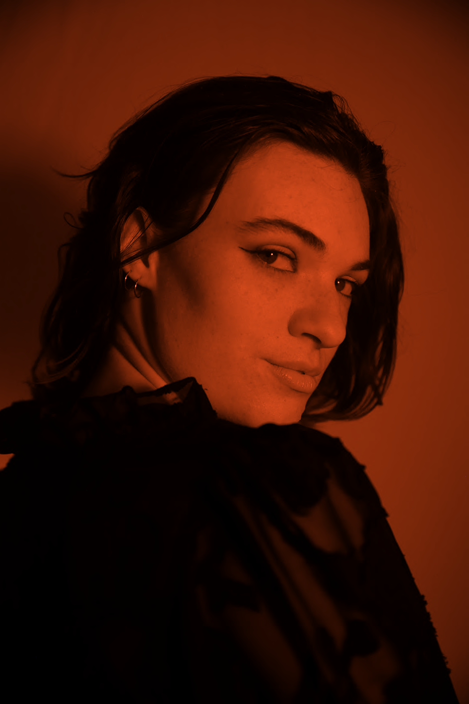 Portrait of a young woman with short dark hair, wearing a black lace top, looking over her shoulder against a warm, reddish background.