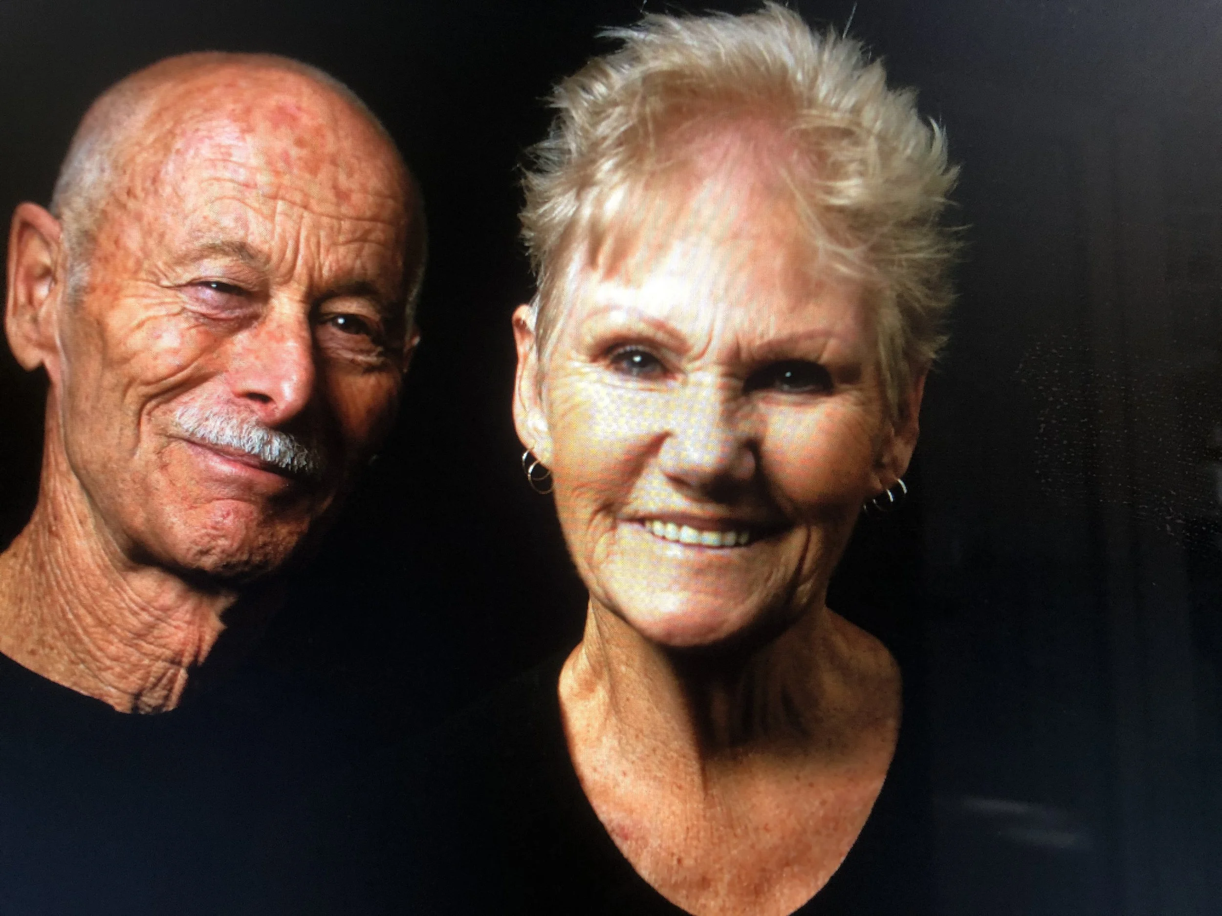Close-up of two elderly individuals, a man and a woman, smiling against a dark background. The man has a bald head with some hair on the sides and wears a slight smile. The woman has short, light-colored, curly hair and is smiling widely, showing her teeth.