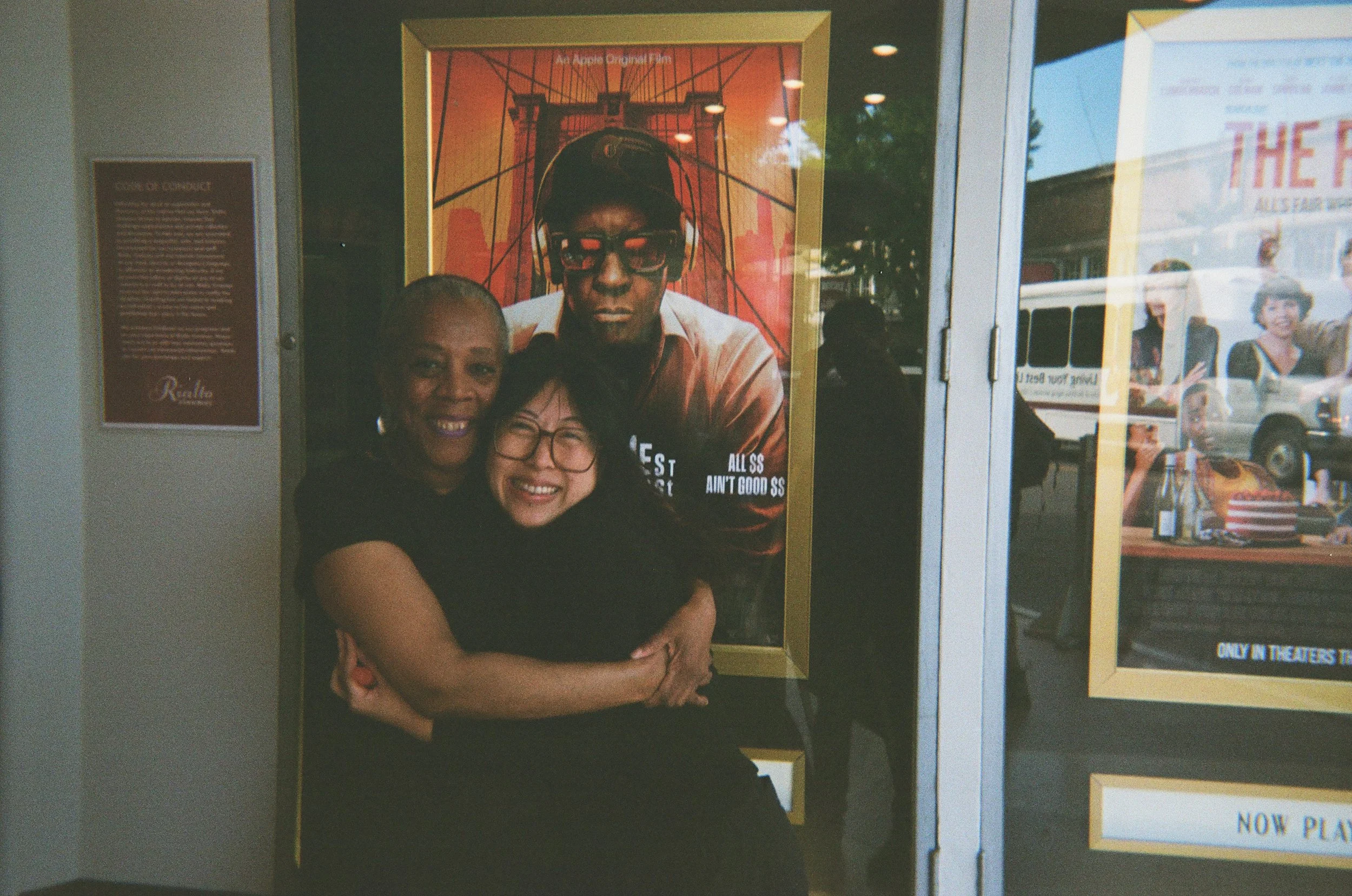 Two movie theater workers hug while standing near a poster of Spike Lee's Highest 2 Lowest, 2025.