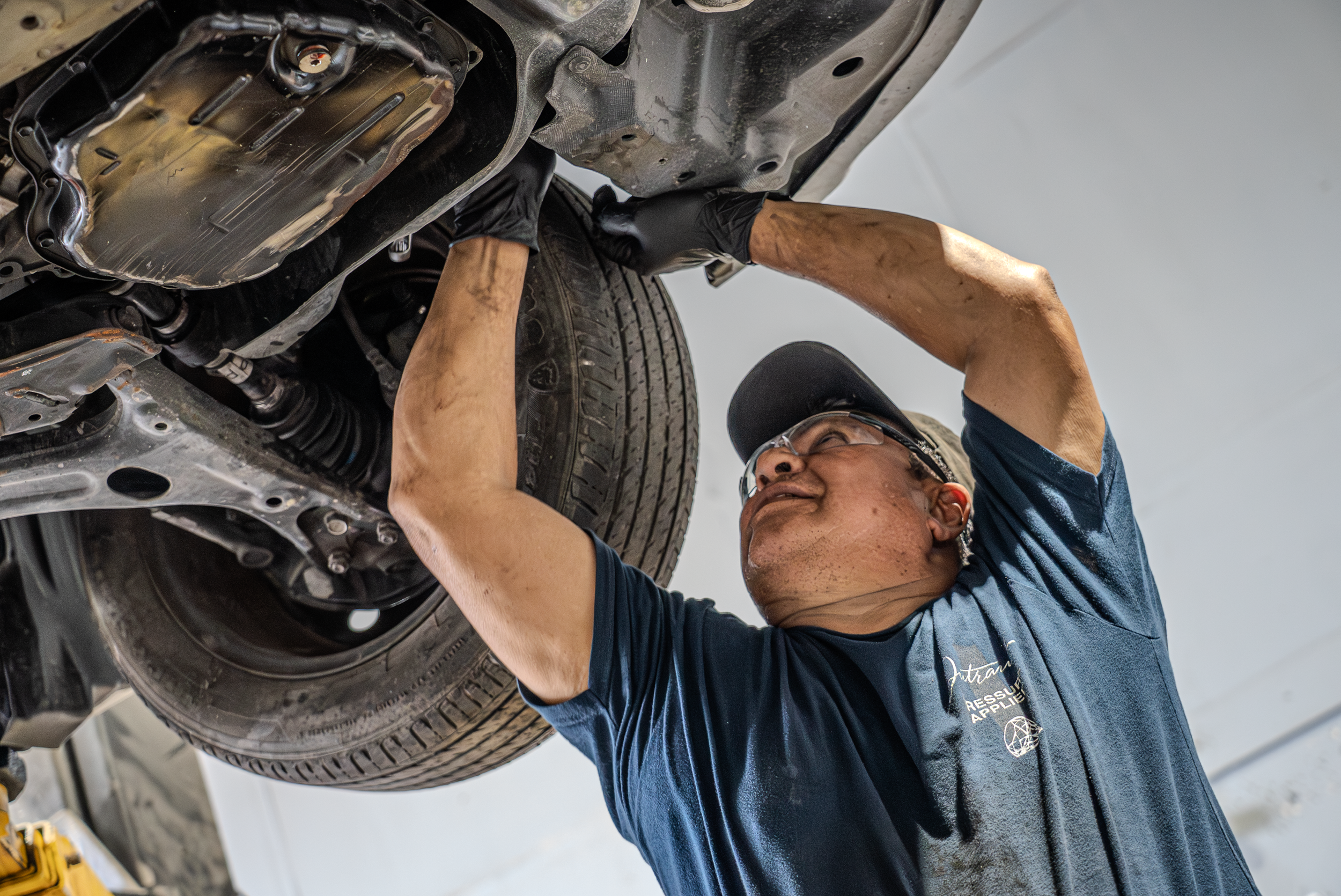 A mechanic working underneath a car lifted on a hydraulic lift, inspecting or repairing the underside of the vehicle.