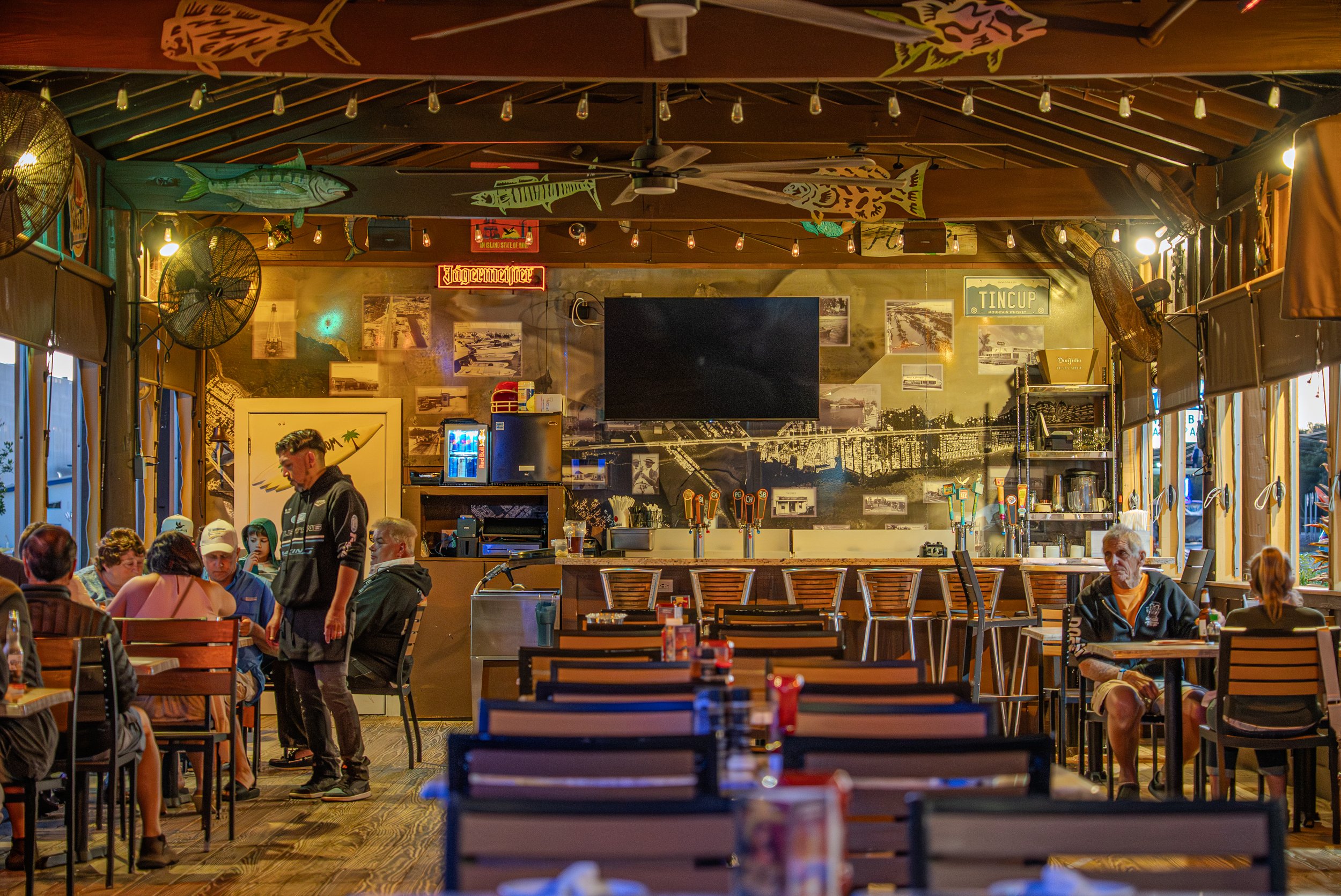 Inside a lively restaurant with wooden floors and tables filled with people dining, decorated with fish and outdoor-themed artwork, string lights, ceiling fans, and a large black TV on the wall.
