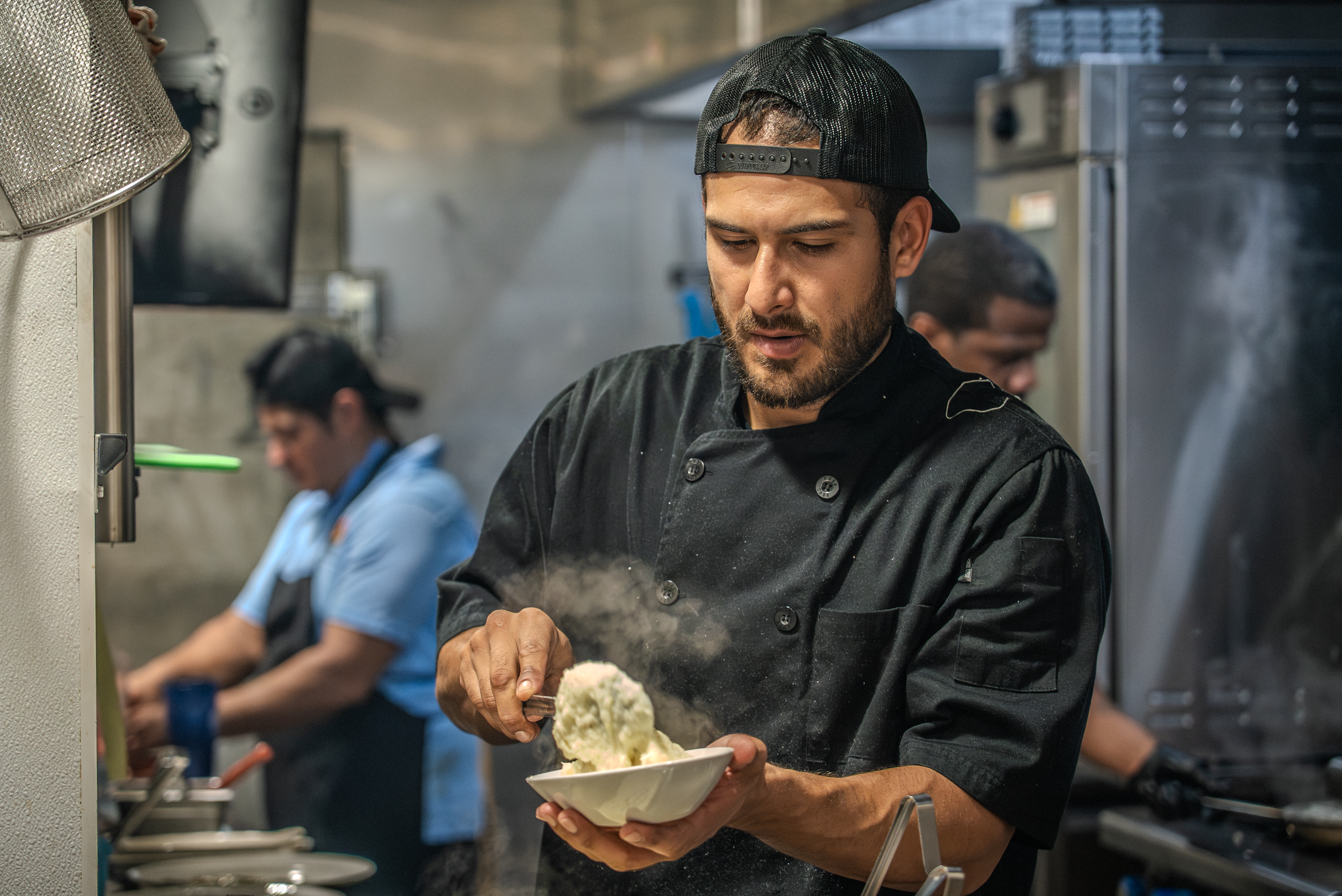 A male chef wearing a black chef jacket and a black mesh cap is serving food into a bowl in a commercial kitchen, with another kitchen worker visible in the background.