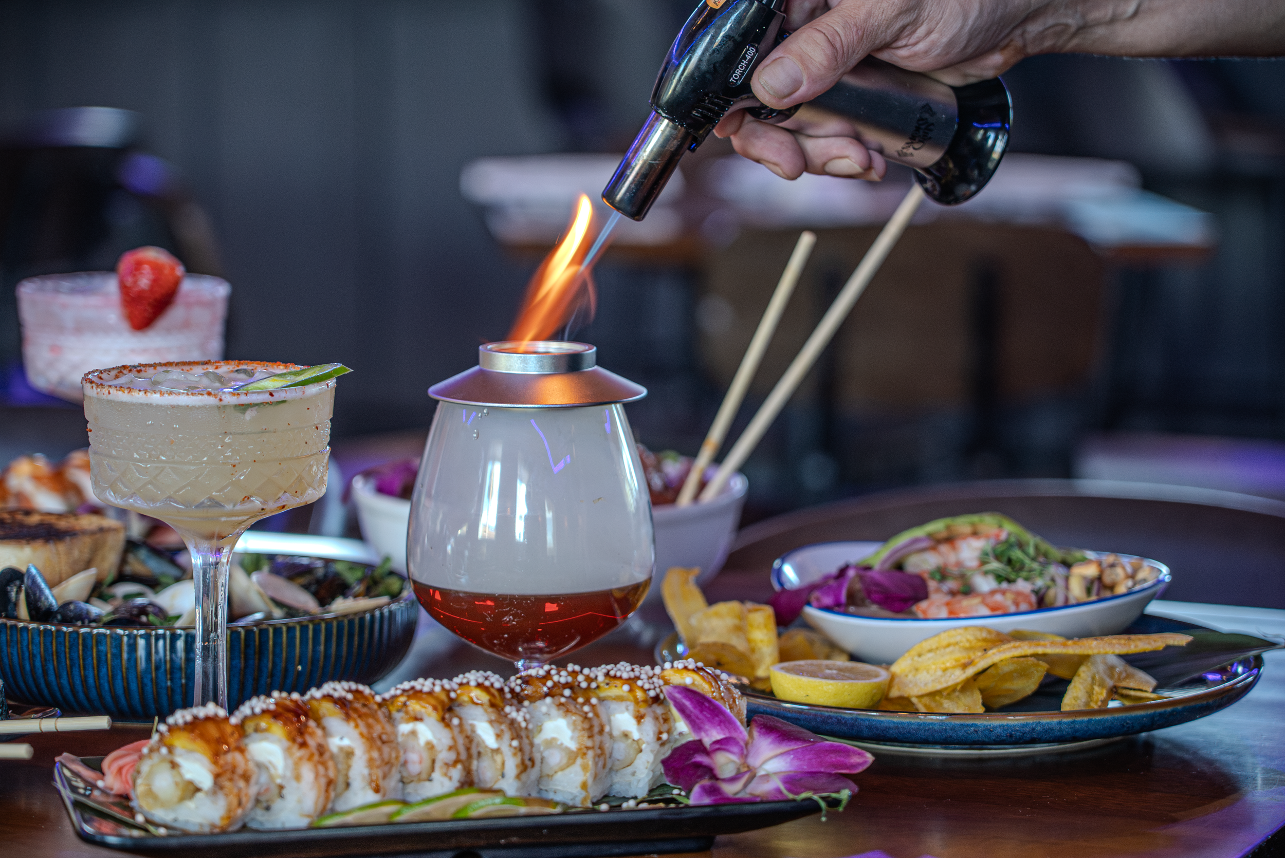 A variety of food dishes including sushi, a shrimp cocktail, salads, lemon slices, and drinks are arranged on a table, with a person's hand using a culinary torch to caramelize or flame a dish.
