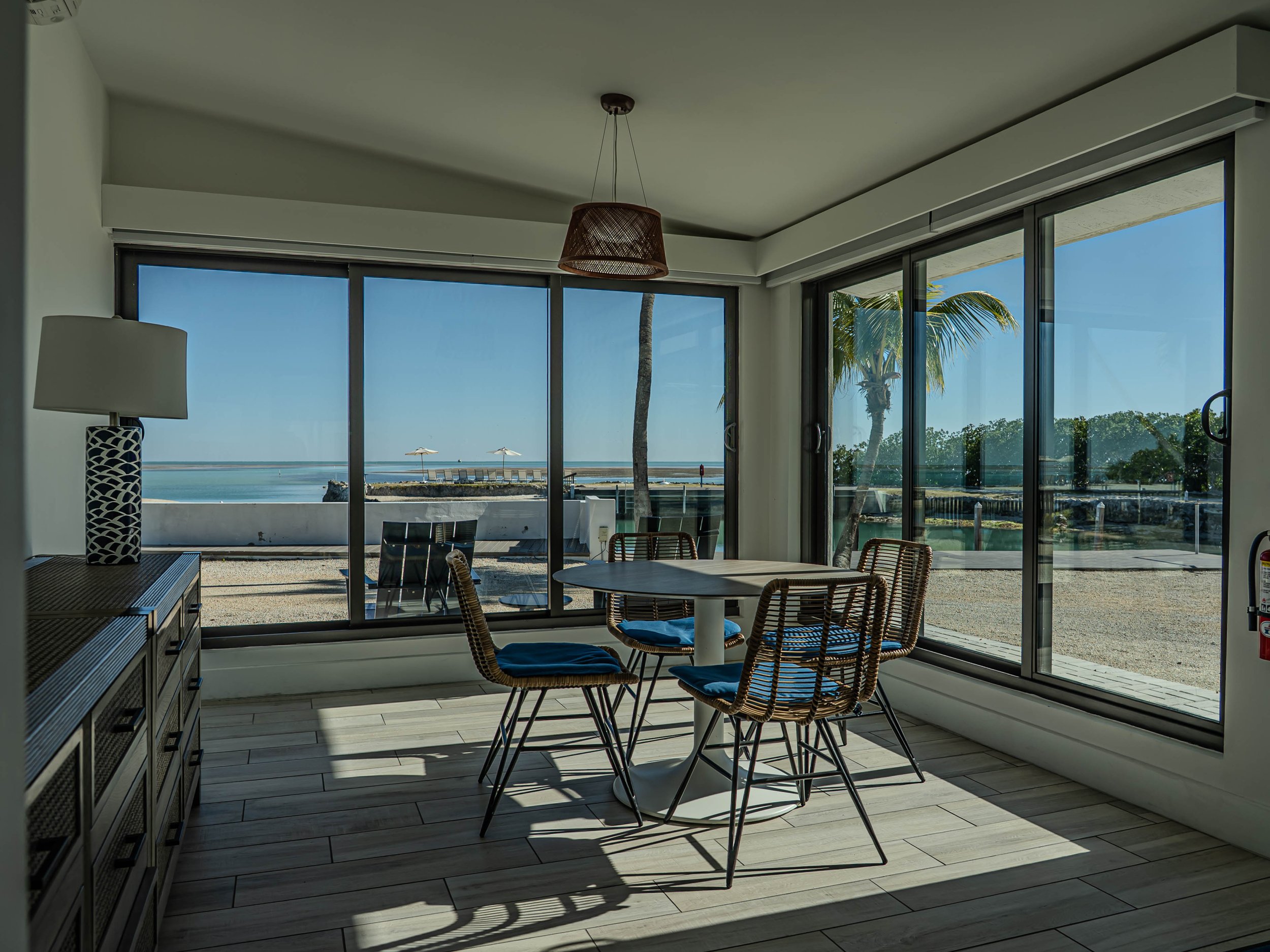 Interior of a beachfront condo with large glass sliding doors open to a view of the ocean, with patio chairs and umbrellas visible outside. The room features a round table with wicker chairs, a tall lamp, and a ceiling light fixture.